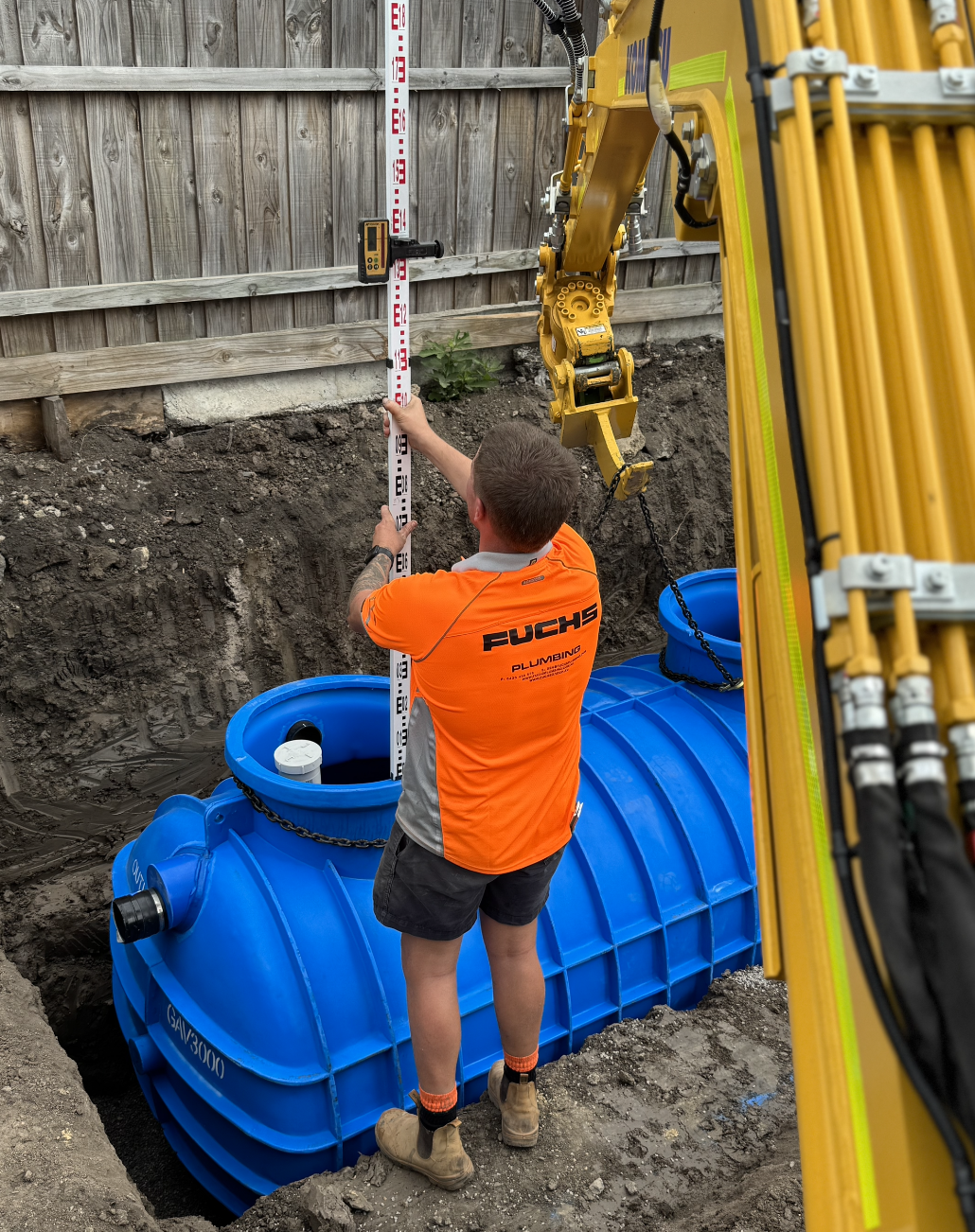 Worker in orange t-shirt measuring a large blue underground water tank being installed in a construction site, with a yellow construction machine and wooden fence in background.