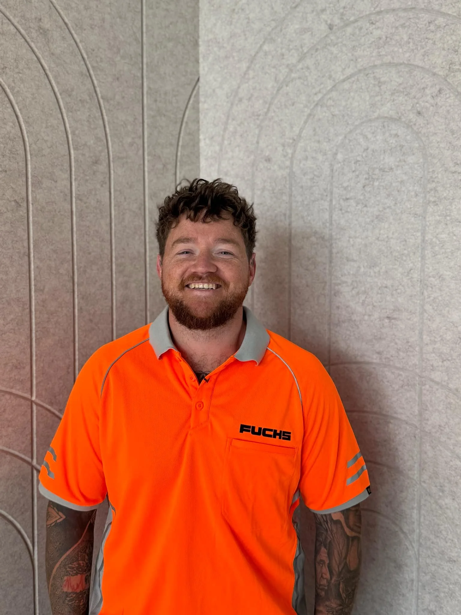 A man with curly hair and a beard, smiling, wearing an orange Fuchs work uniform shirt with grey accents, standing against a textured wall with geometric patterns.