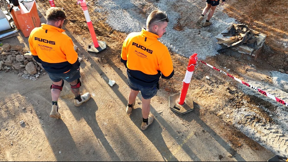 Two construction workers in orange shirts working on a road, with uneven ground, safety cones, and barrier tape nearby.