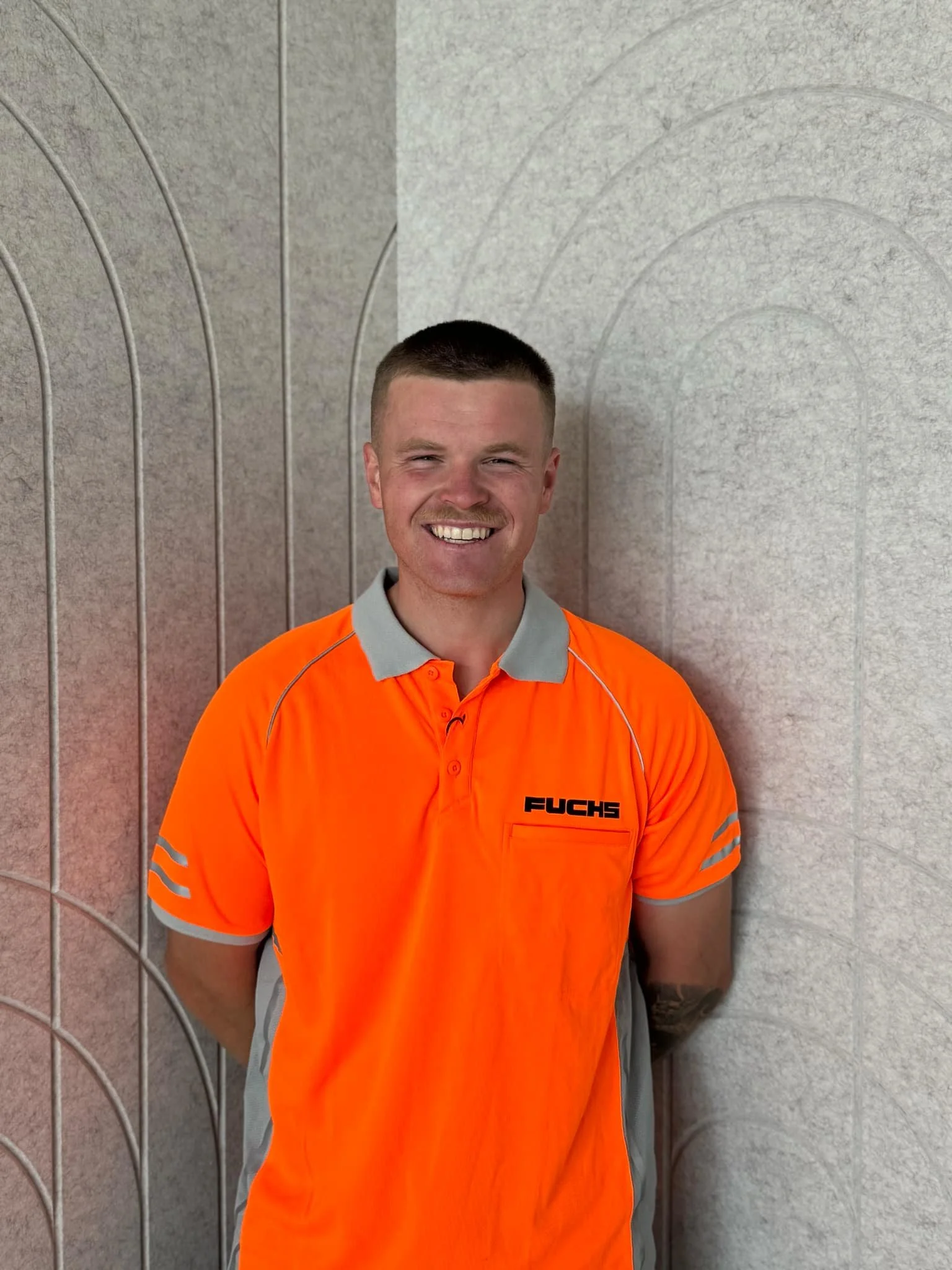 A young man with short hair, smiling, wearing an orange work shirt with gray accents and the brand name "Fuchs" on it, standing beside a textured wall with arch patterns.