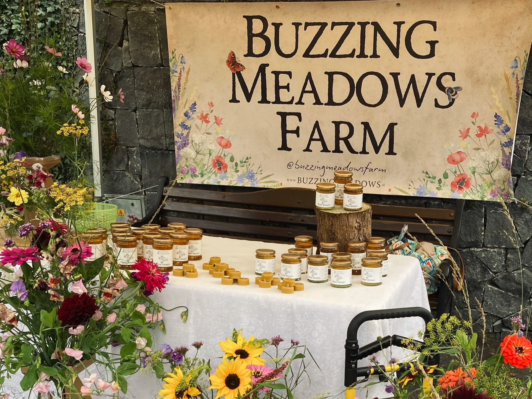 A display table at Buzzing Meadows Farm selling jars of honey, with a large banner in the background and colorful flowers surrounding the table.
