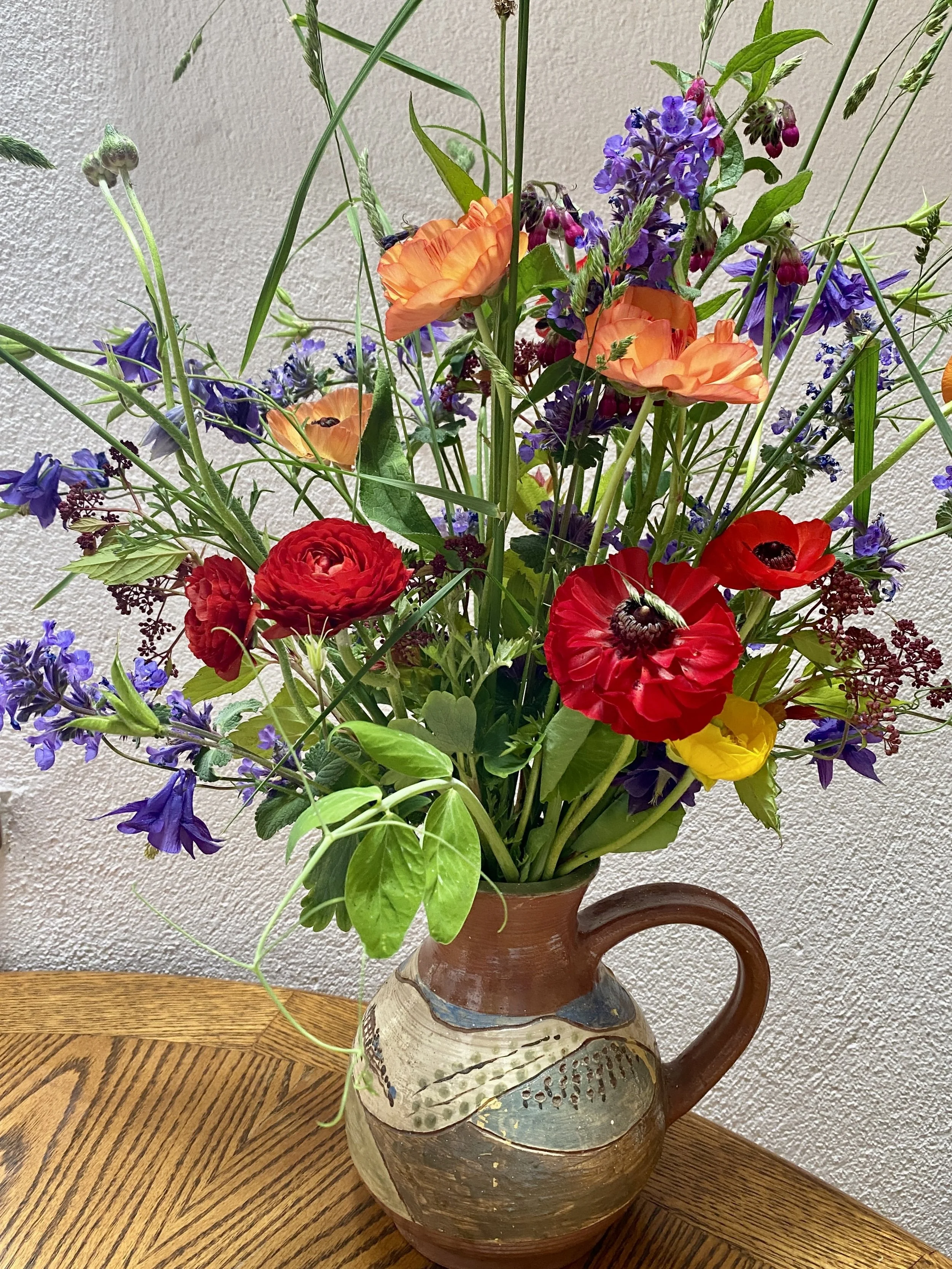 Colorful bouquet of flowers in a brown ceramic vase on a wooden table against a textured beige wall.