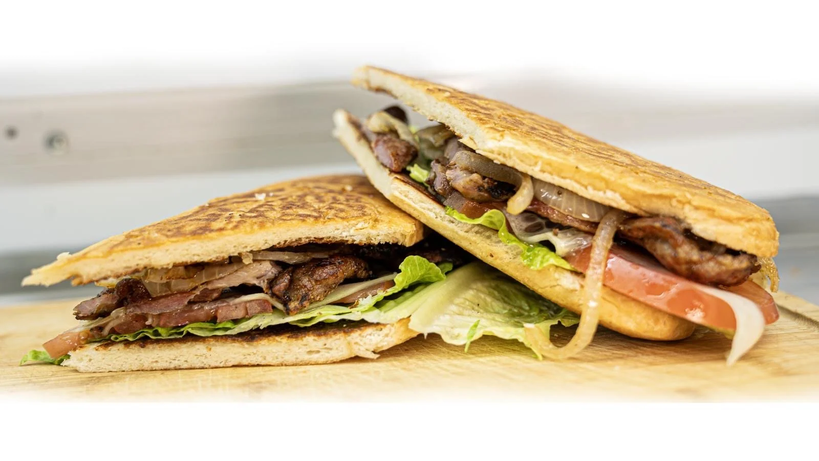 Two grilled sandwiches with lettuce, tomato, onions, bacon, sausage, and cheese on whole wheat bread placed side by side on a wooden surface.