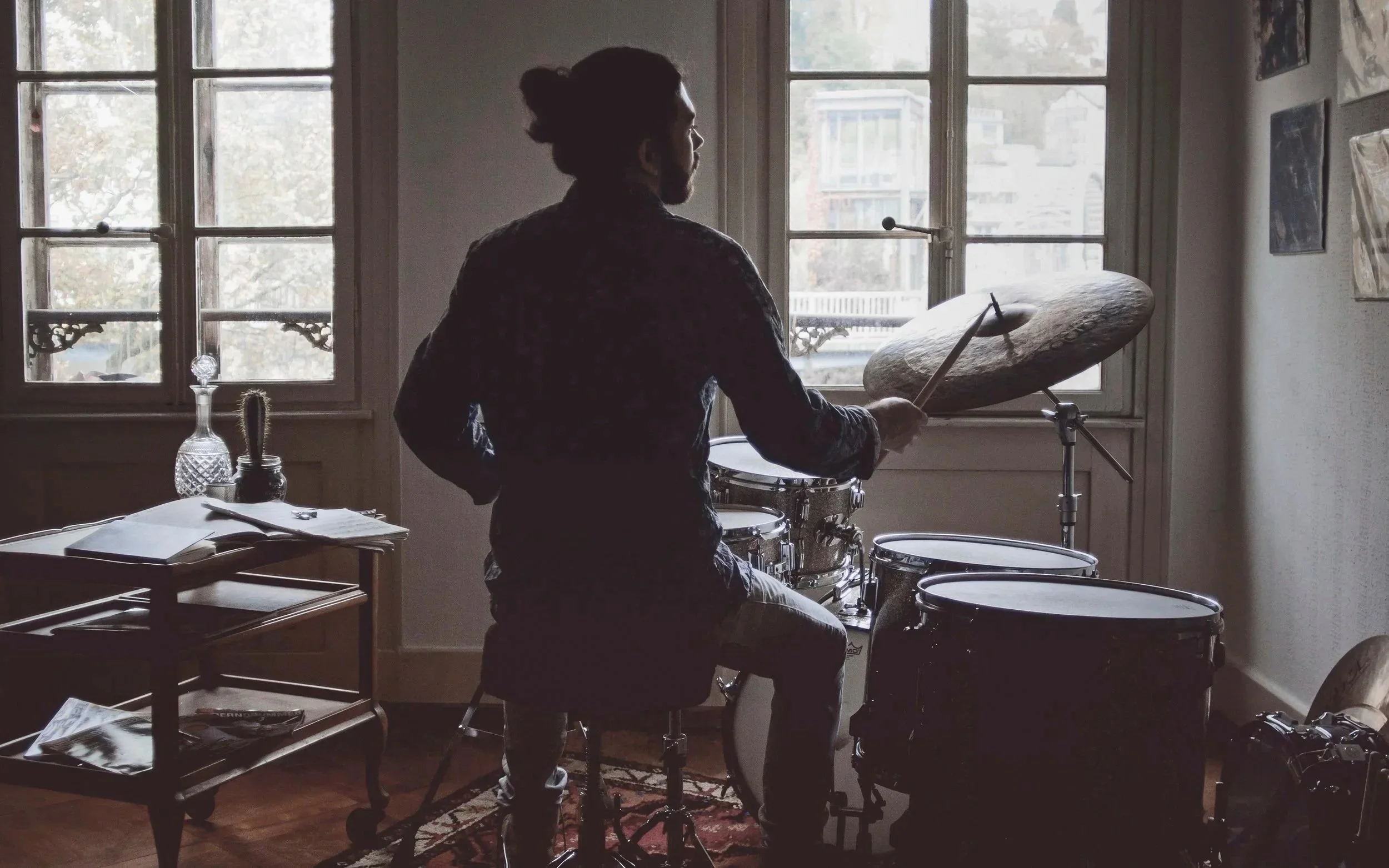 A silhouette of a person with long hair in a bun sitting on a drum throne and playing a drum set near large windows in a well-lit room.