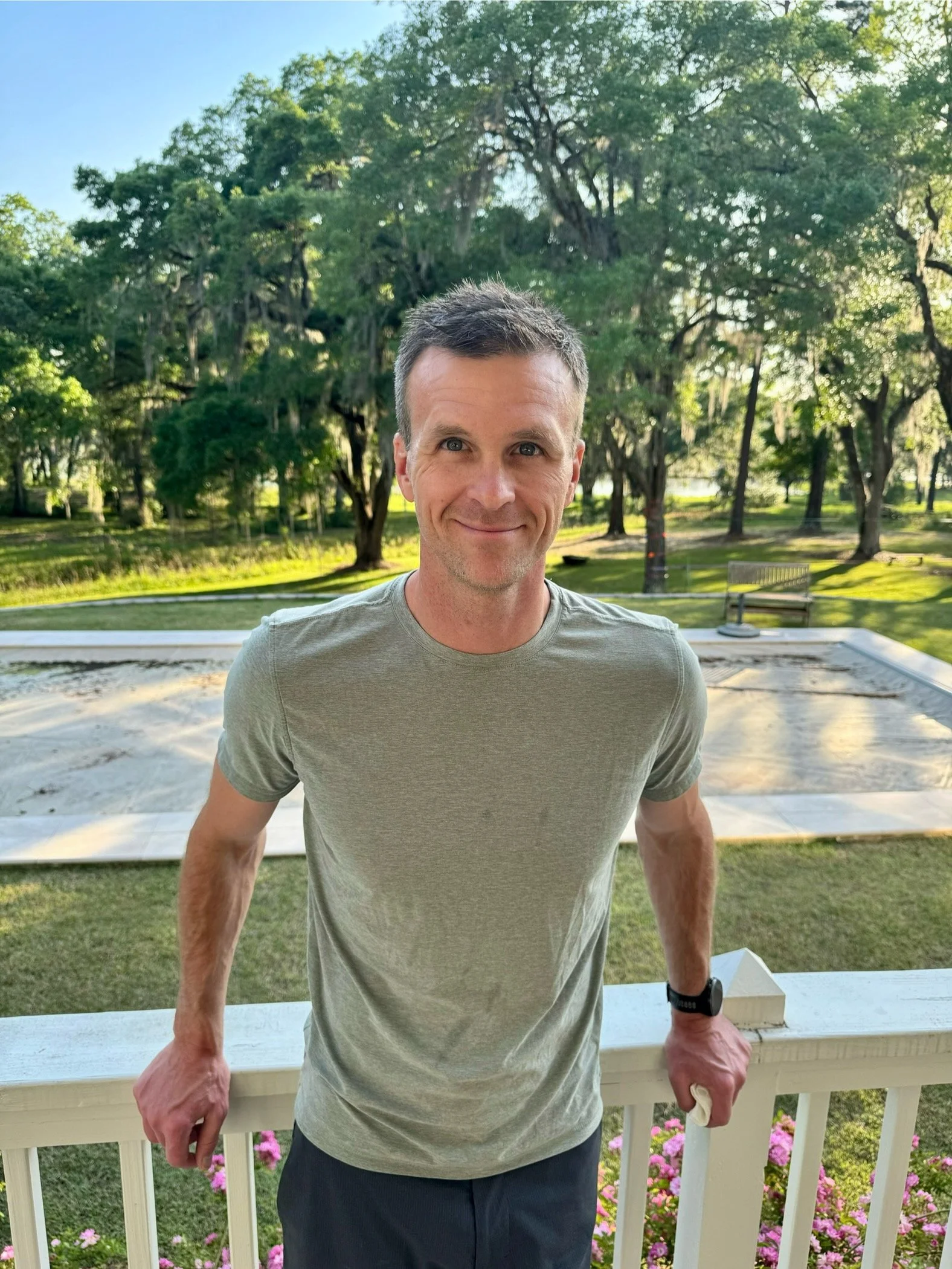 A man wearing a green t-shirt, smiling, leaning back on a ledge with a a beautiful tree-scape in the background.