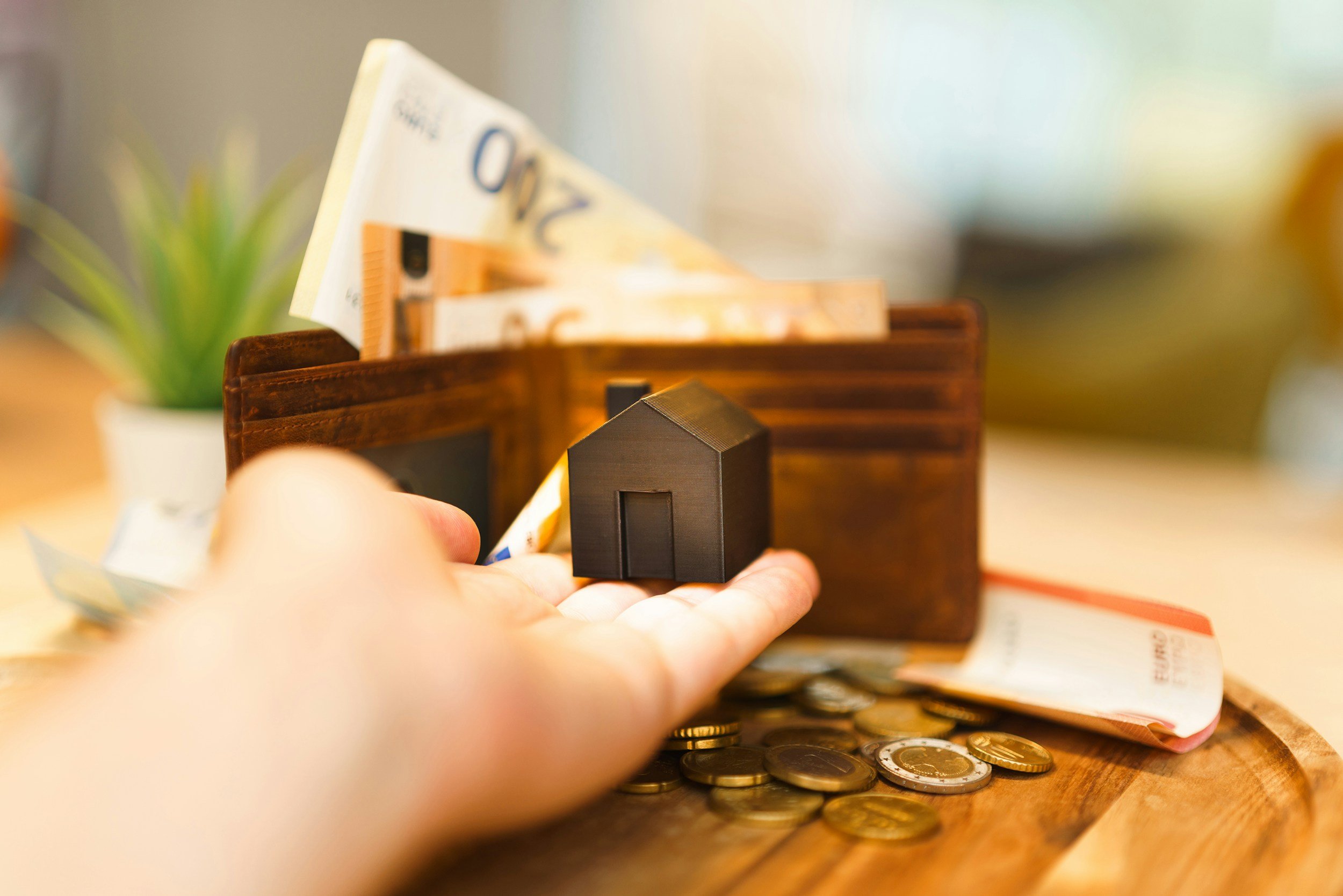 A mans hand holding an animated miniture home in front of a wallet with money all around.