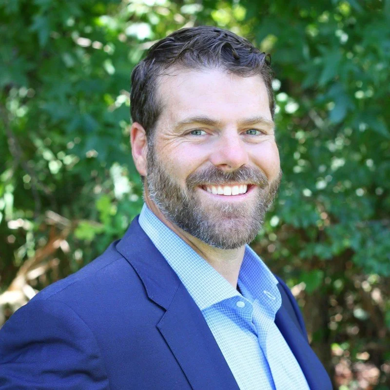 Headshot of a man with brown hair and a beard, smiling, wearing a blue blazer and light blue dress shirt, outdoors with green foliage in the background.