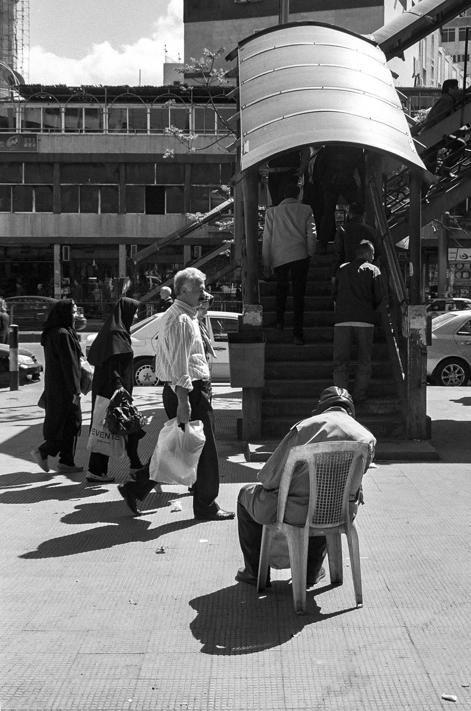 People walking up stairs through an outdoor staircase in an urban setting, with parked cars and a building in the background. A man is sitting on a plastic chair in the foreground.