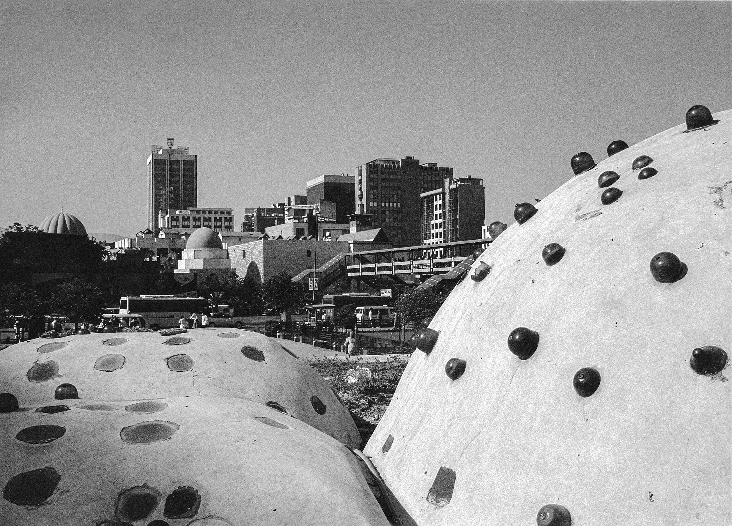 Black and white photo of a city skyline with modern high-rise buildings and domed structures in the foreground, including large, rounded concrete shapes with embedded metal bolts. Damascus Syria-Hussein Haddad