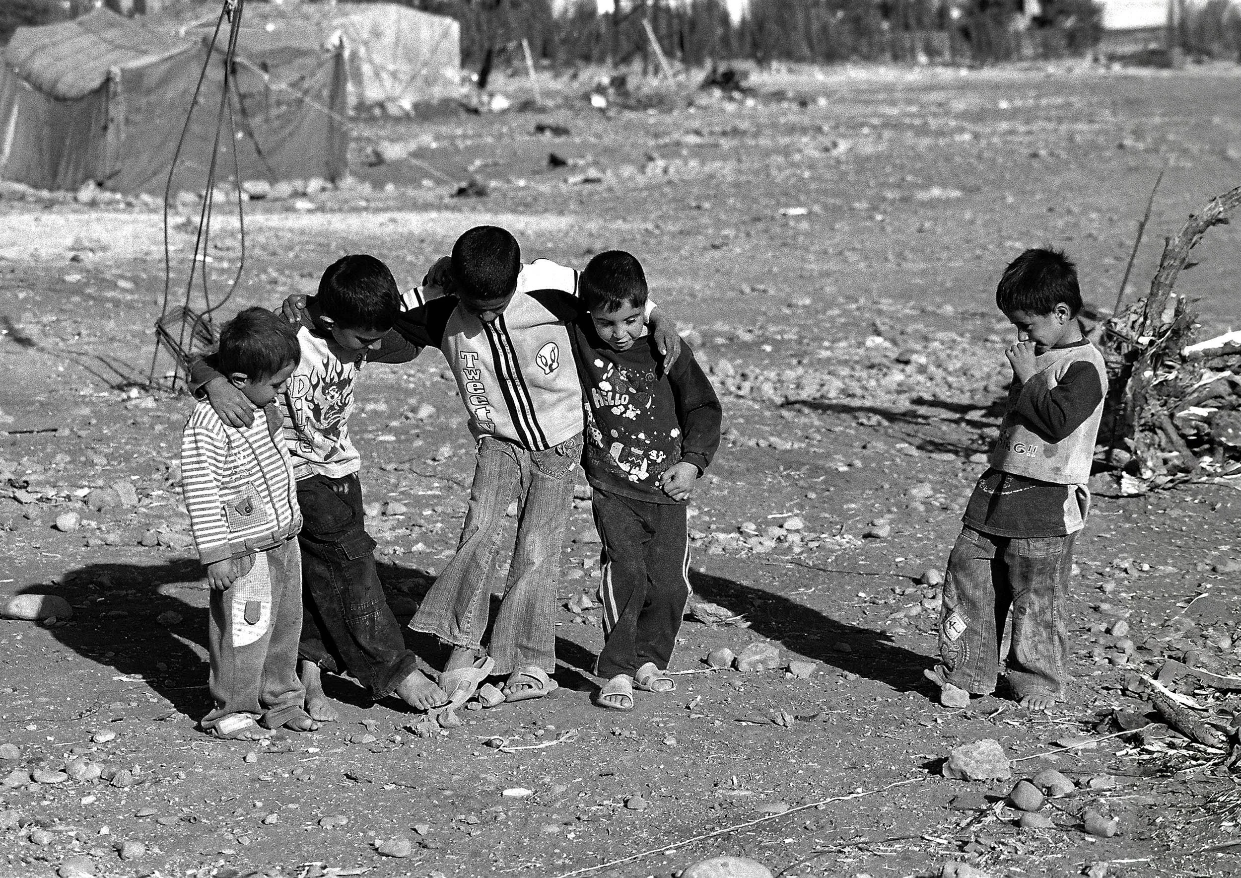 Five young boys standing on rocky ground, with arms around each other, appearing to be looking down at something on the ground. One boy on the right is standing apart, looking down and touching his face. The background shows a barren landscape with s