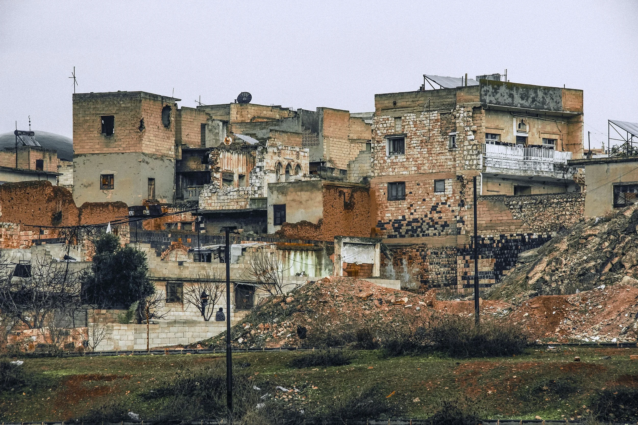 A hillside neighborhood with multiple weathered buildings made of brick and concrete, some with visible damage, set against a cloudy sky.