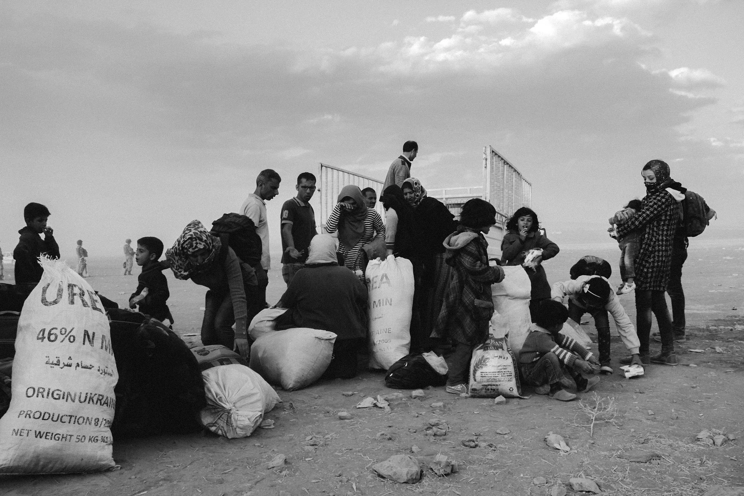 Group of people, including women and children, gather outdoors with bags and belongings, with a metal structure and cloudy sky in the background.