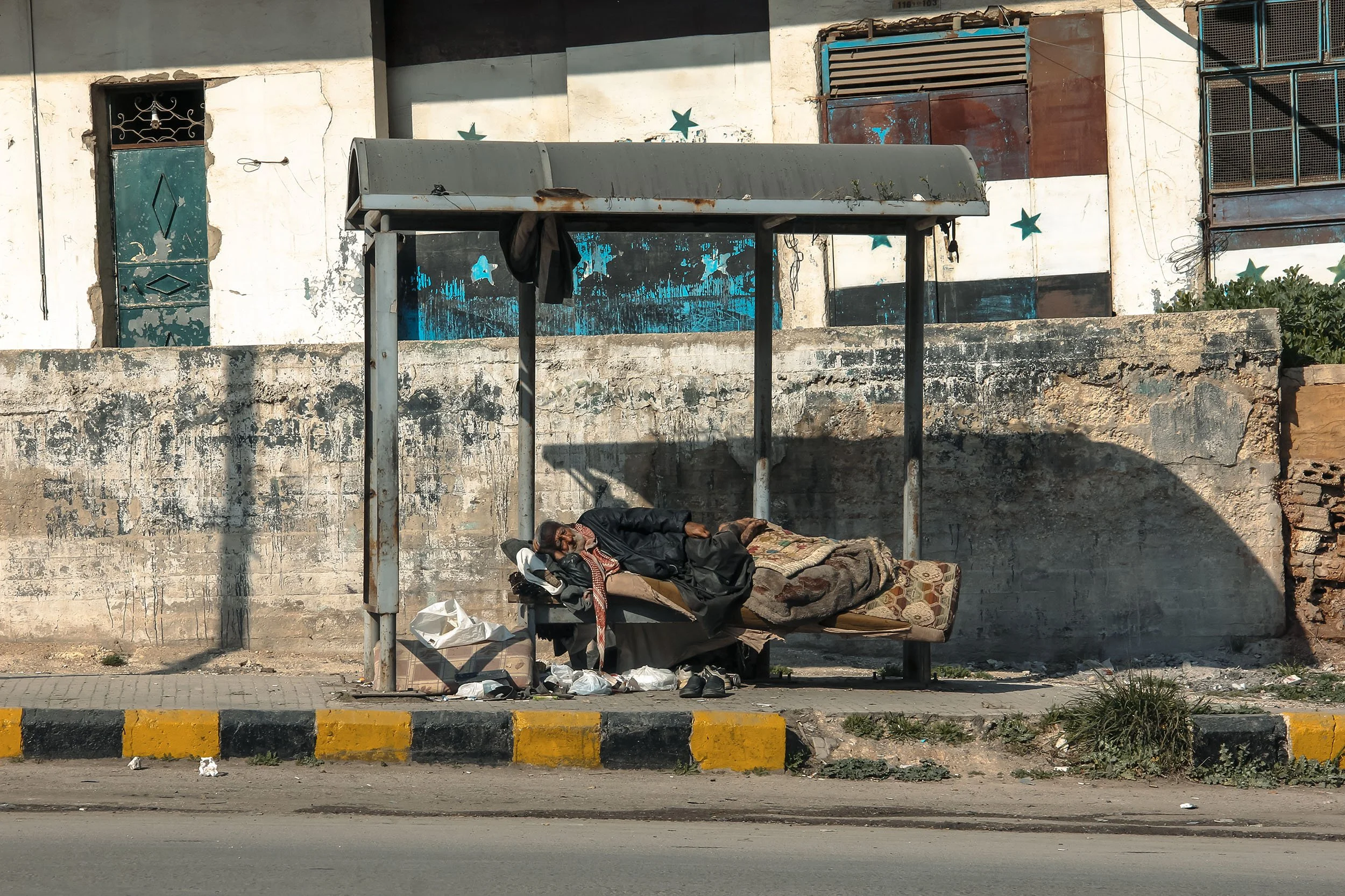 A person sleeping on a bench under a bus shelter on an urban street. The person is covered with blankets and has various belongings around them. The background shows a weathered wall with painted designs and a building with windows.