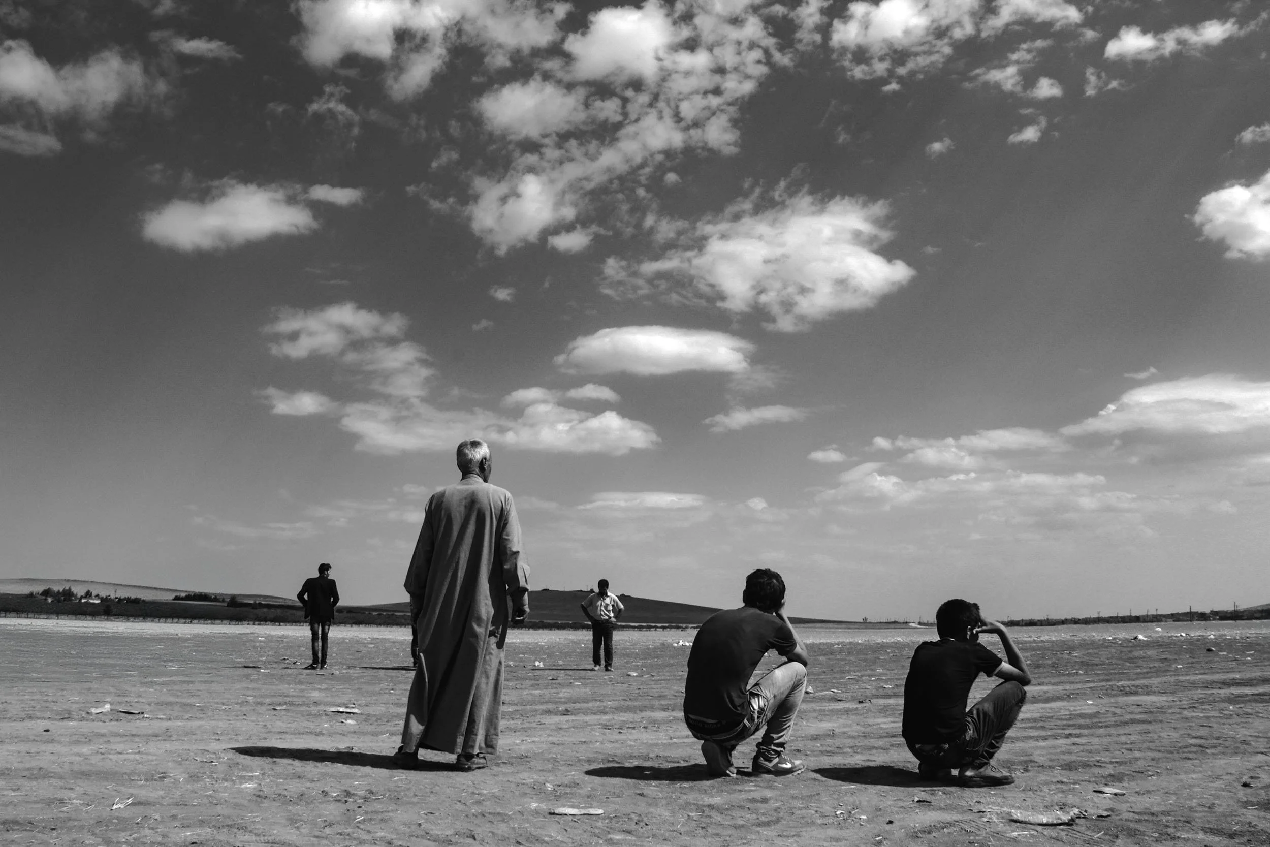 Five men on a vast, open landscape, two are sitting, one is standing, and two are walking in the distance, under a sky filled with clouds