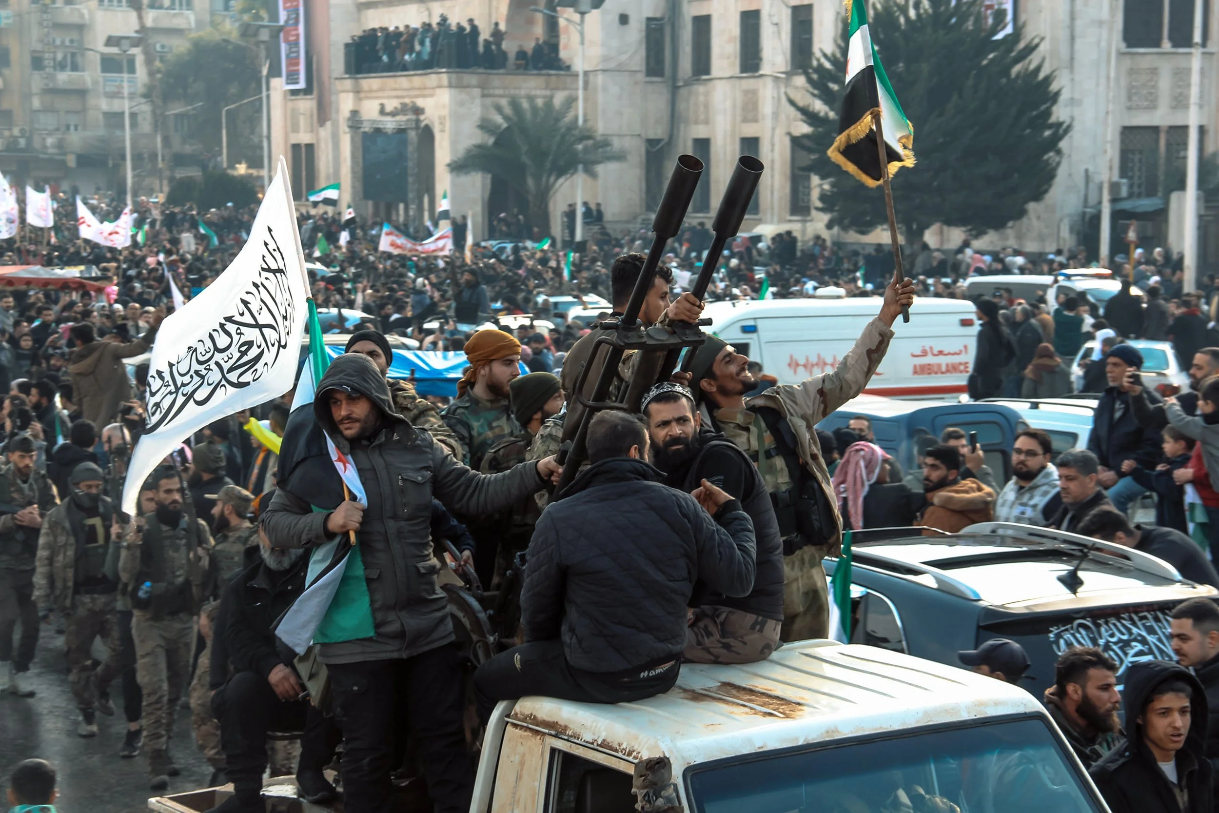A large crowd of protesters and fighters on and around a white pick-up truck in a city street, holding flags, including a white flag with Arabic script and green and black flags, with many bystanders and vehicles, and a building with balconies in the