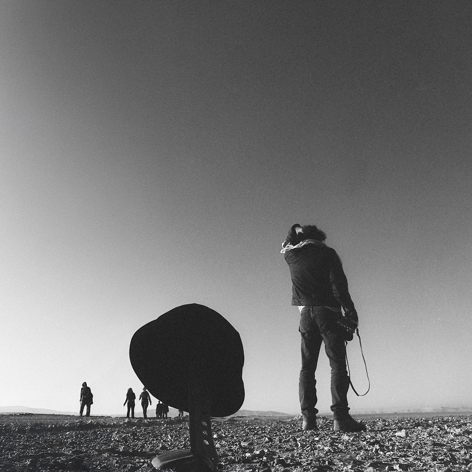Black and white photo of a person standing on a beach with their head turned and hand on their head, holding a camera. Four people are in the background near the horizon, and a hat is in the foreground on the sandy ground.