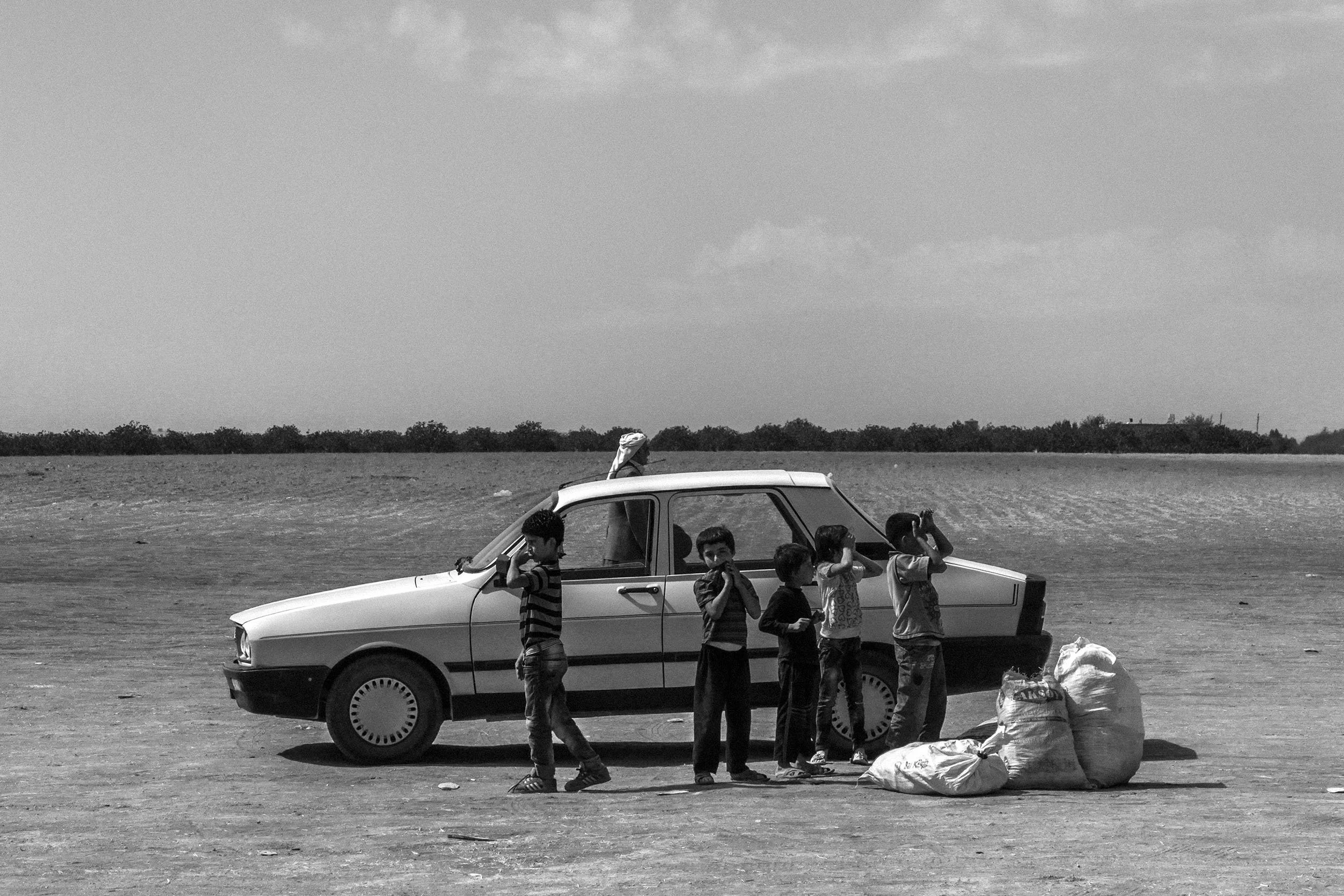 Six children standing and leaning against a car on a barren landscape, with large bags on the ground nearby, under a cloudy sky.