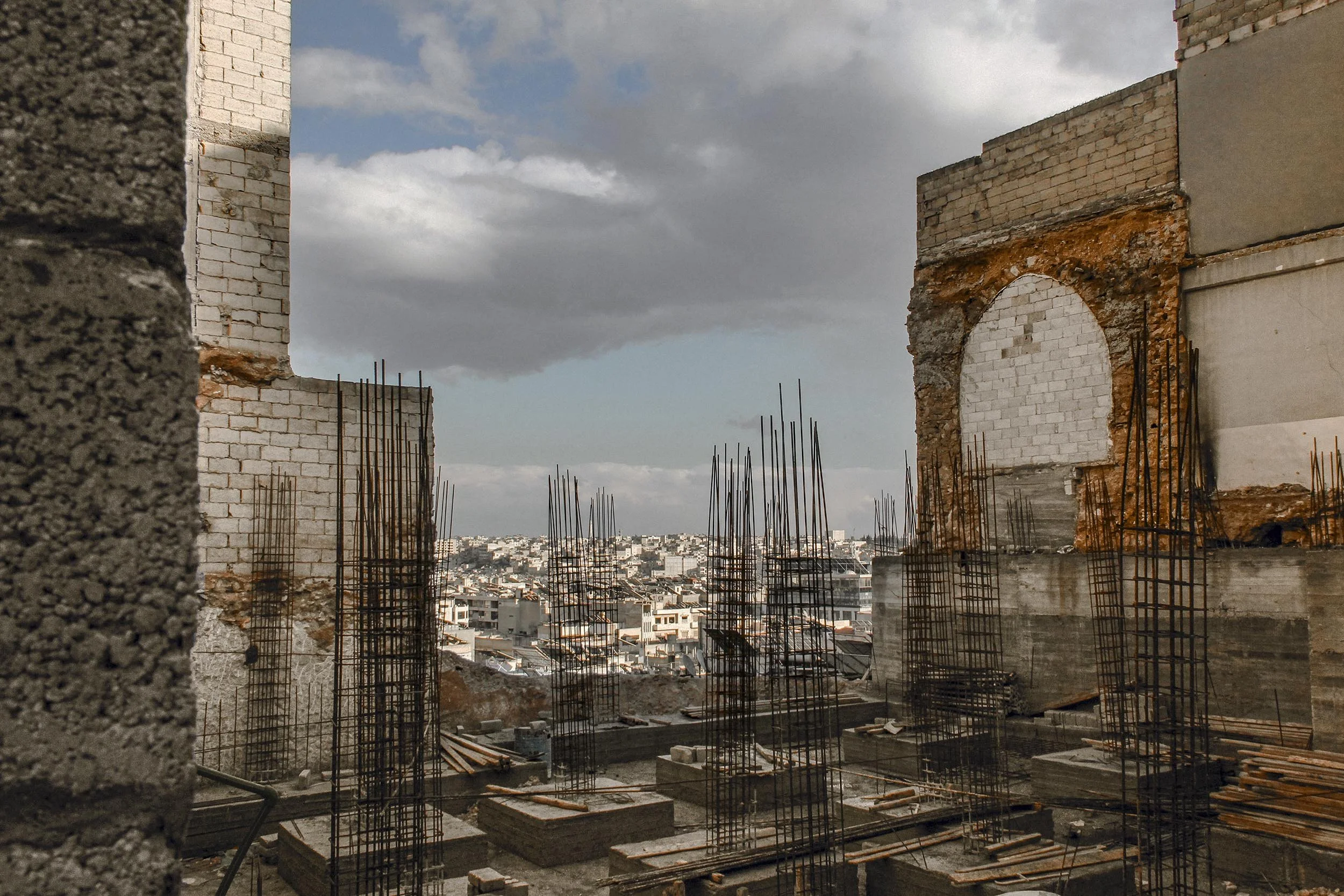 Construction site with brick walls and metal rebar; city skyline in the background under a partly cloudy sky.