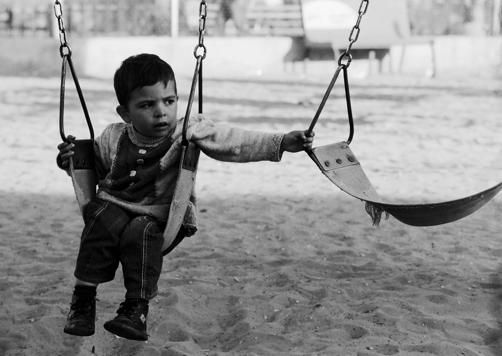 A young boy sitting alone on a swing in a playground, looking to the side with a serious expression, in black and white.Damascus Syria-Hussein Haddad