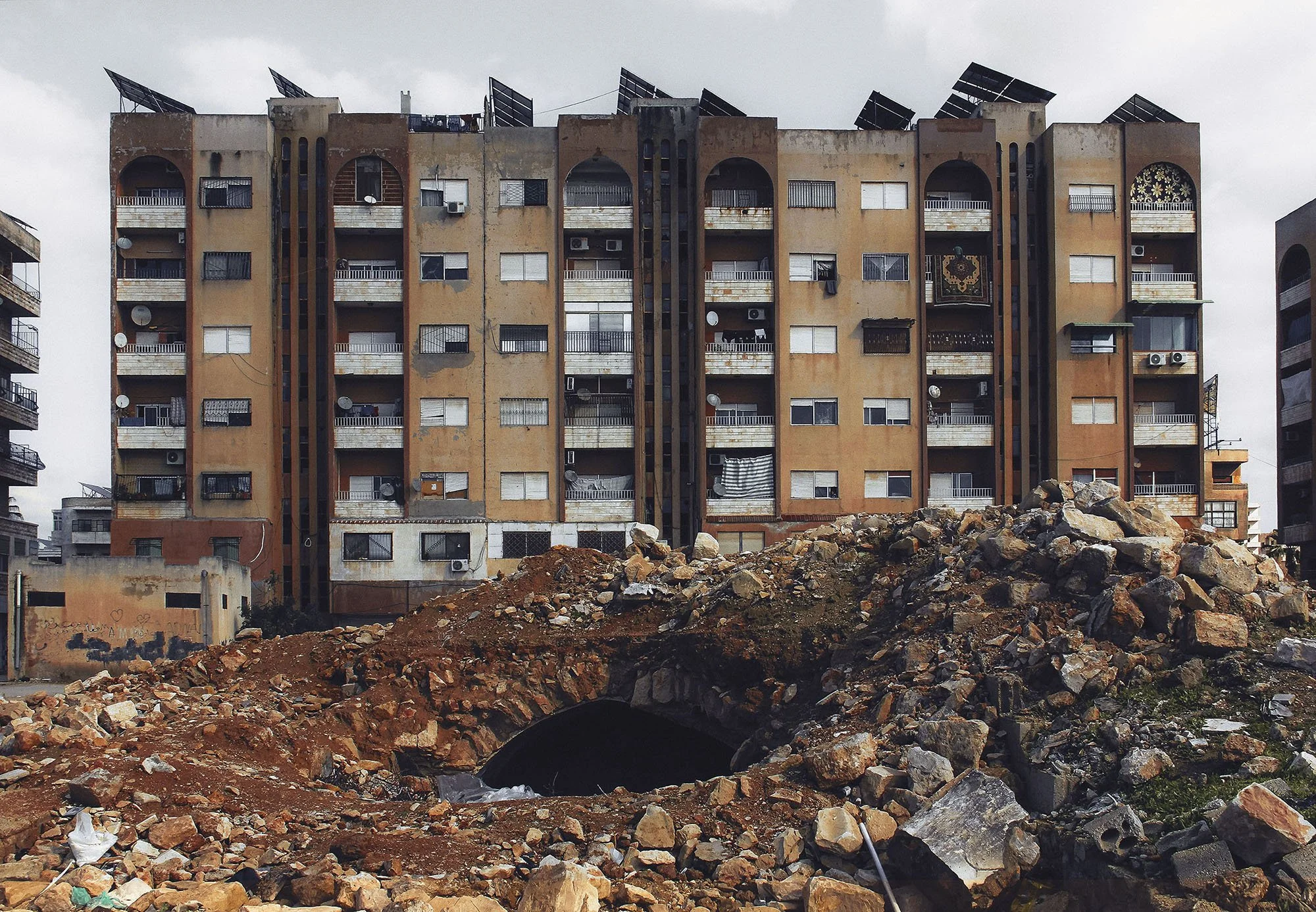 A multi-story apartment building with beige and brown exterior, multiple balconies, and solar panels on the roof. In the foreground, there is a large pile of rocks and dirt, with an underground tunnel opening and some debris, suggesting construction 