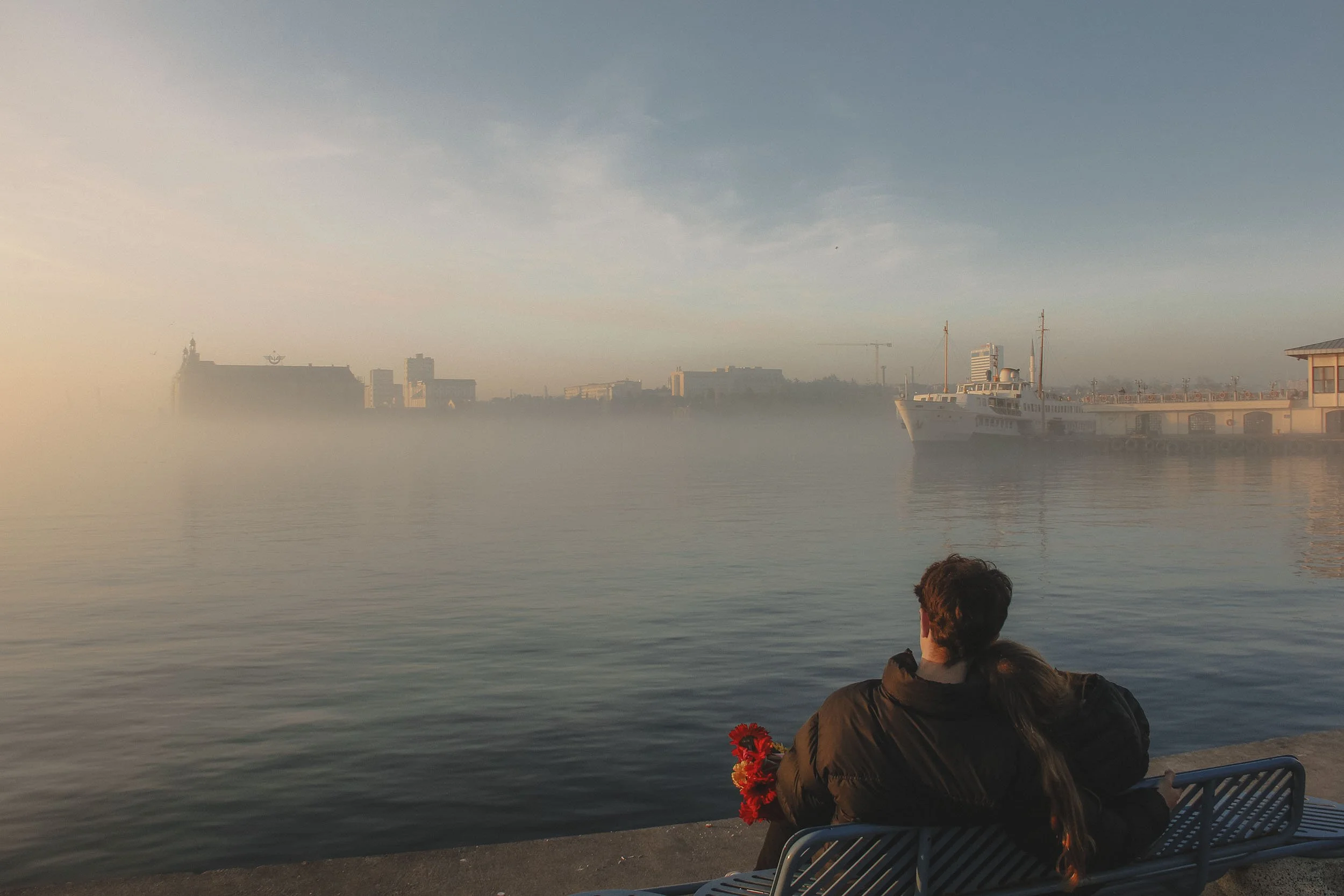 A couple sitting on a bench by the water during sunrise, holding flowers, with a foggy city skyline and a large boat in the background.