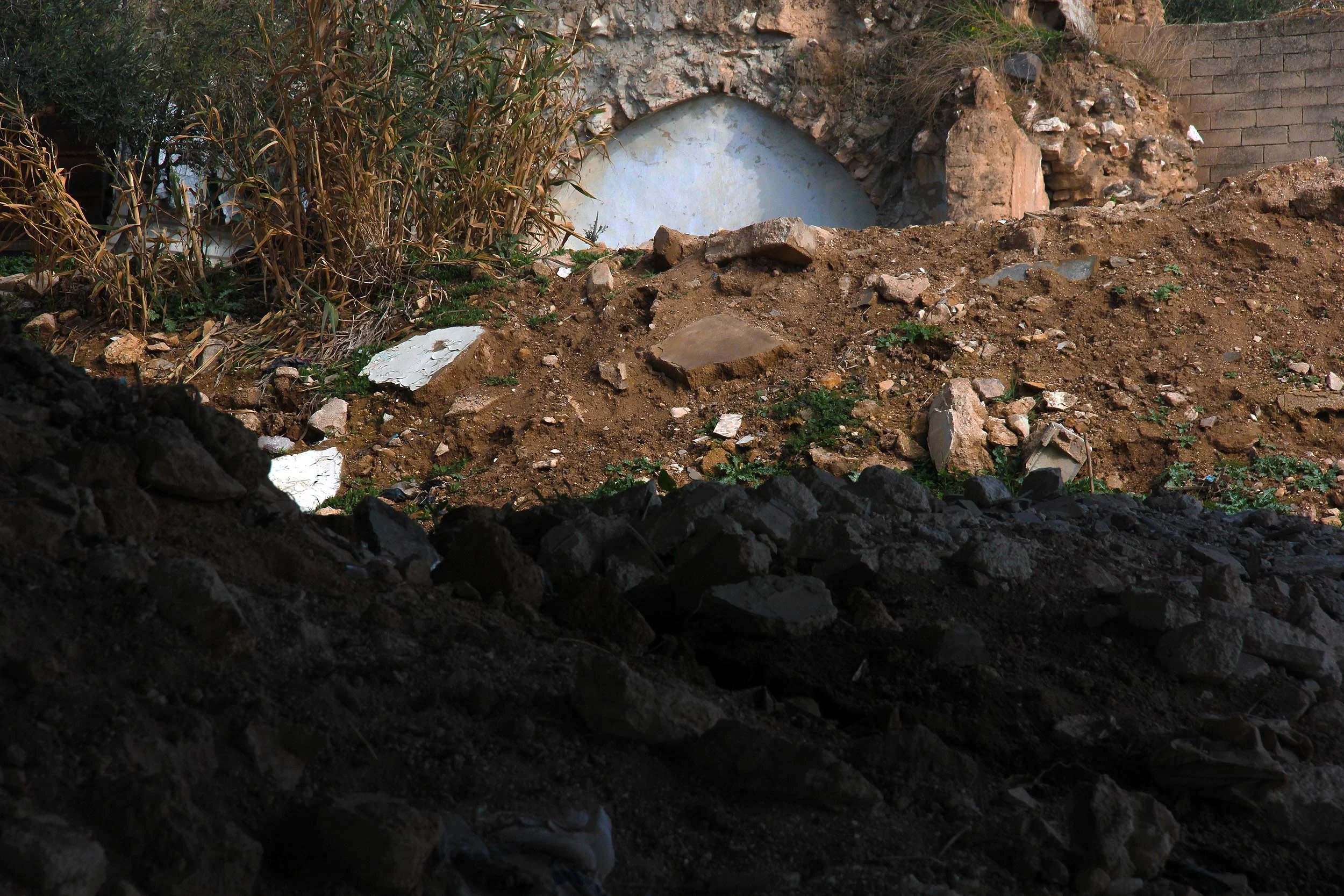 A dirt and rock-filled excavation site with dried plants and debris, and an arched concrete structure in the background.