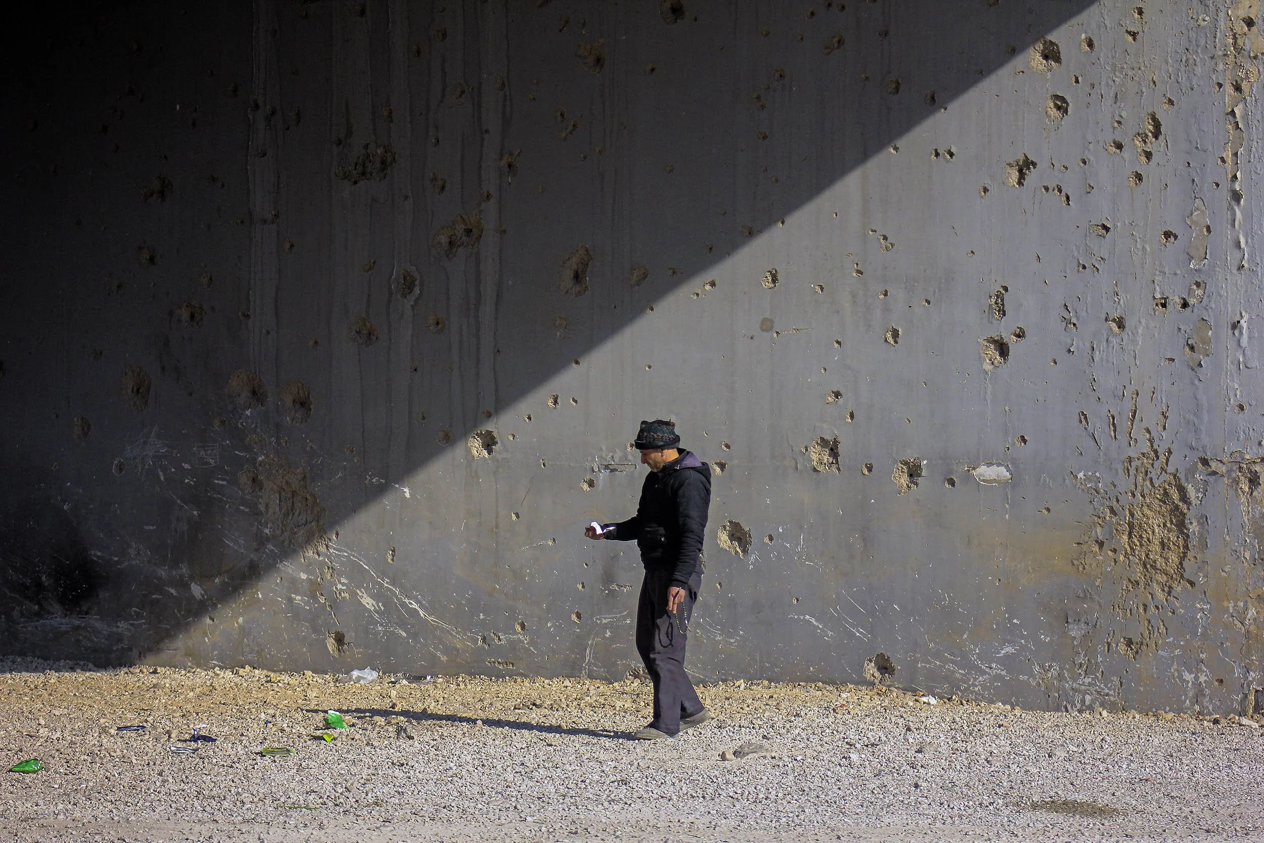 A man dressed in dark clothing, wearing a beanie, standing on gravel ground, looking at a cell phone against a large, weathered concrete wall with numerous holes and patches, with a diagonal shadow across the wall.