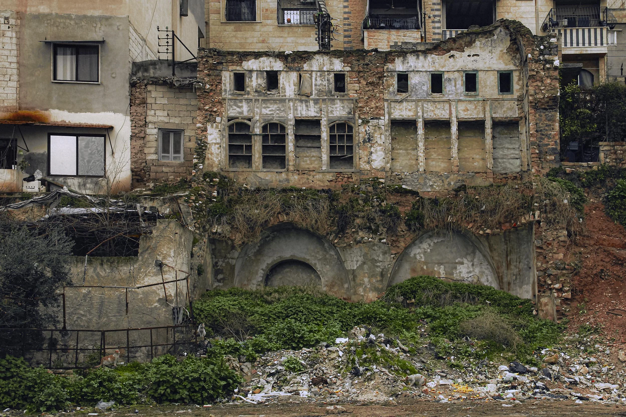 Dilapidated building on a hillside with broken windows, overgrown vegetation, and scattered trash at the base.