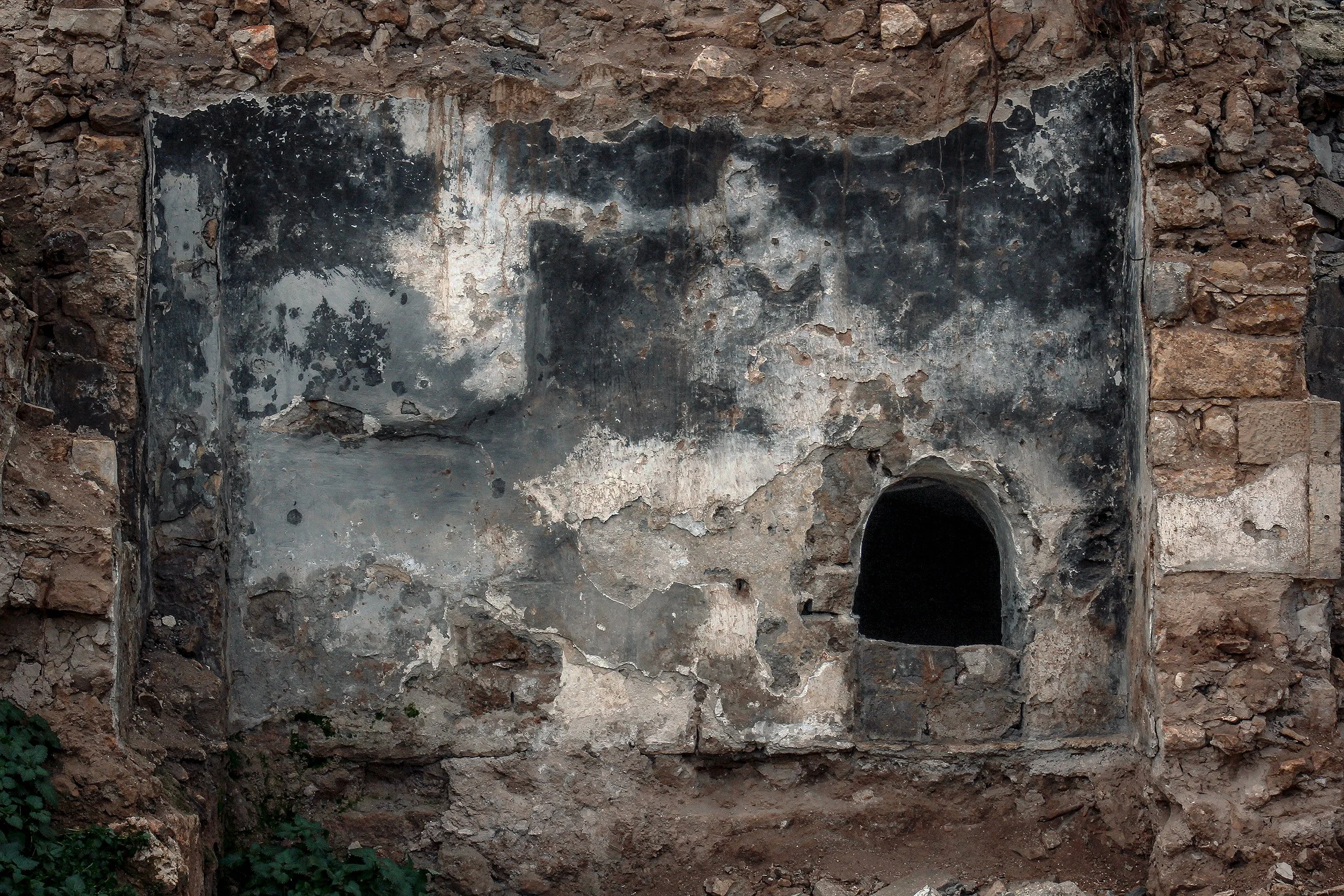 An ancient stone structure with a dark, hollowed-out archway and a partially blackened wall, surrounded by weathered stone bricks.