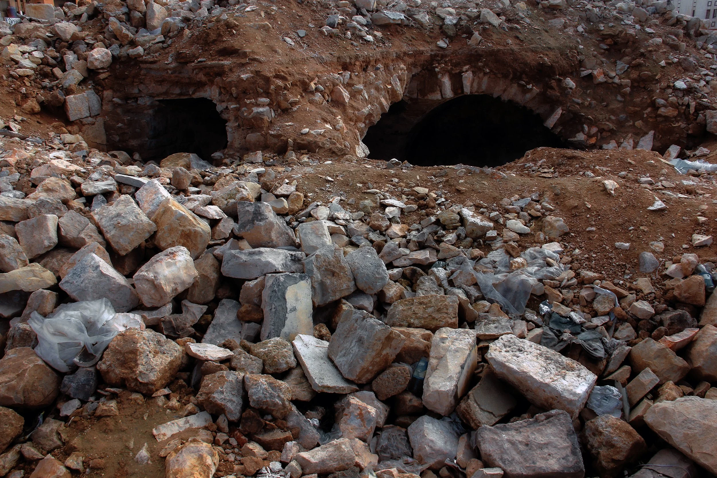 Construction site with two dark tunnel entrances in a dirt hillside, surrounded by scattered rocks and debris.