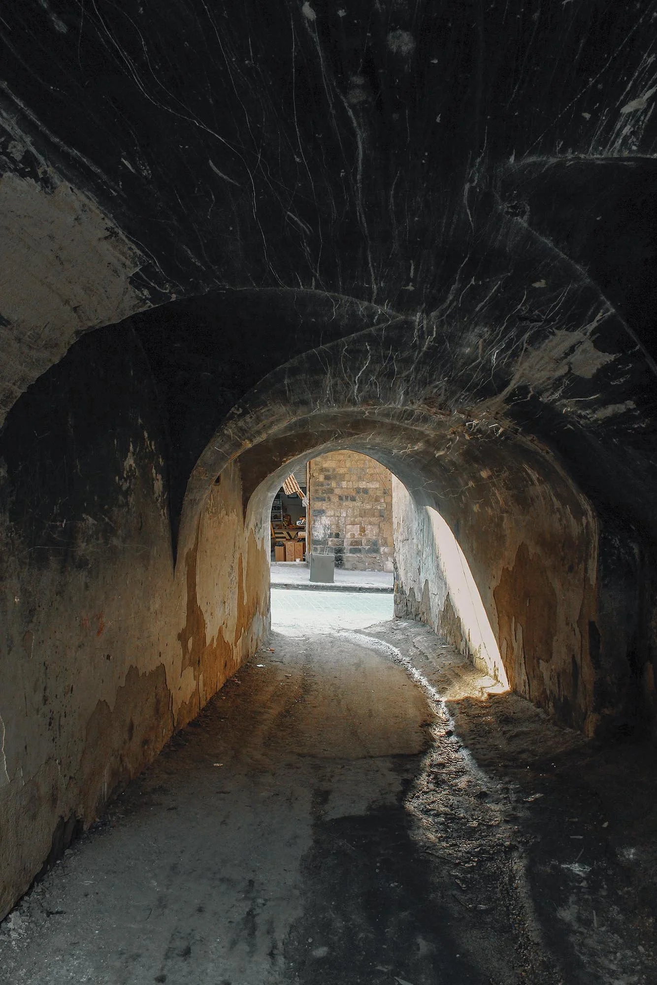 View from inside a dark tunnel with a curved ceiling, leading to a lit exterior with a stone building and a sidewalk.