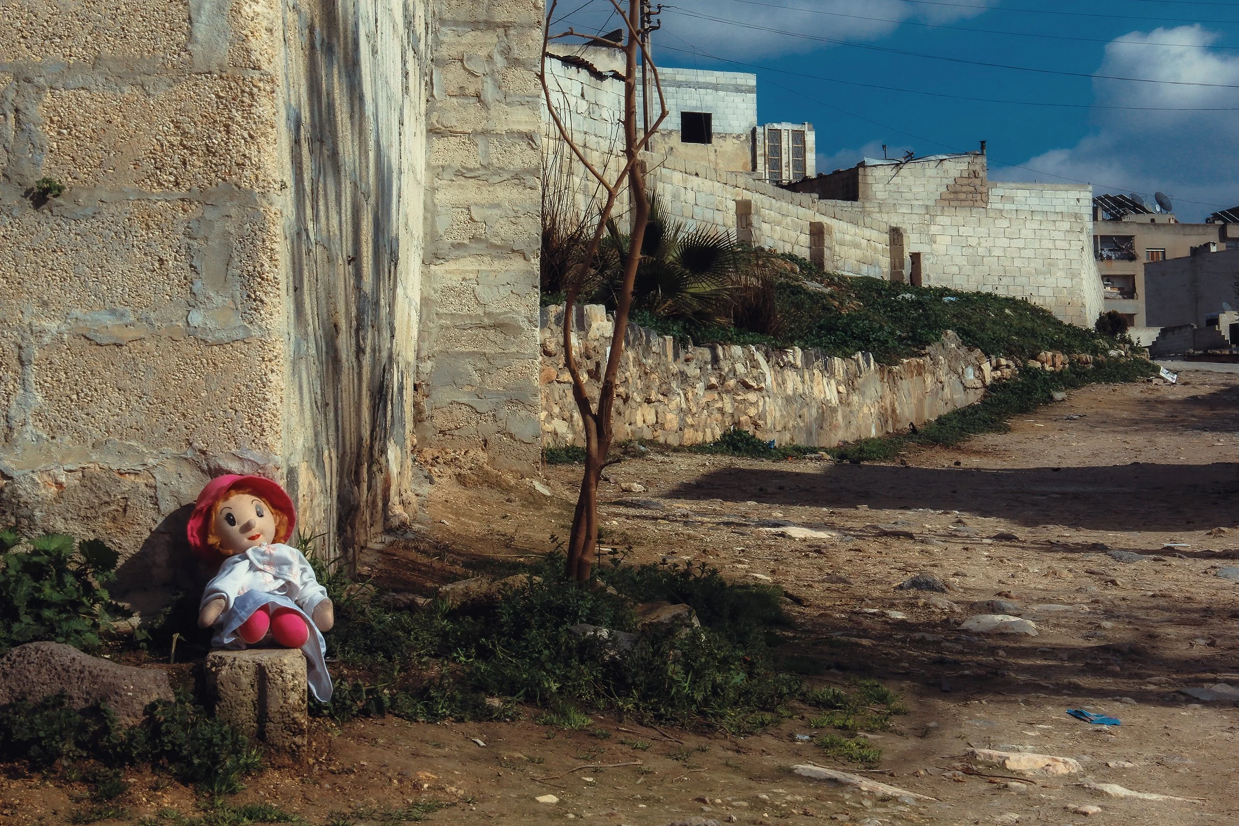 A doll with red hair, white dress, and pink shoes sitting against a stone wall on a dirt ground with a small tree nearby, in a rural area with white buildings and a cloudy sky in the background.