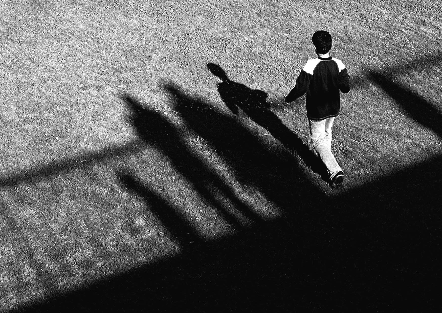 A person walking on a gravel surface with strong shadow patterns cast by a nearby structure, seen from above in black and white.