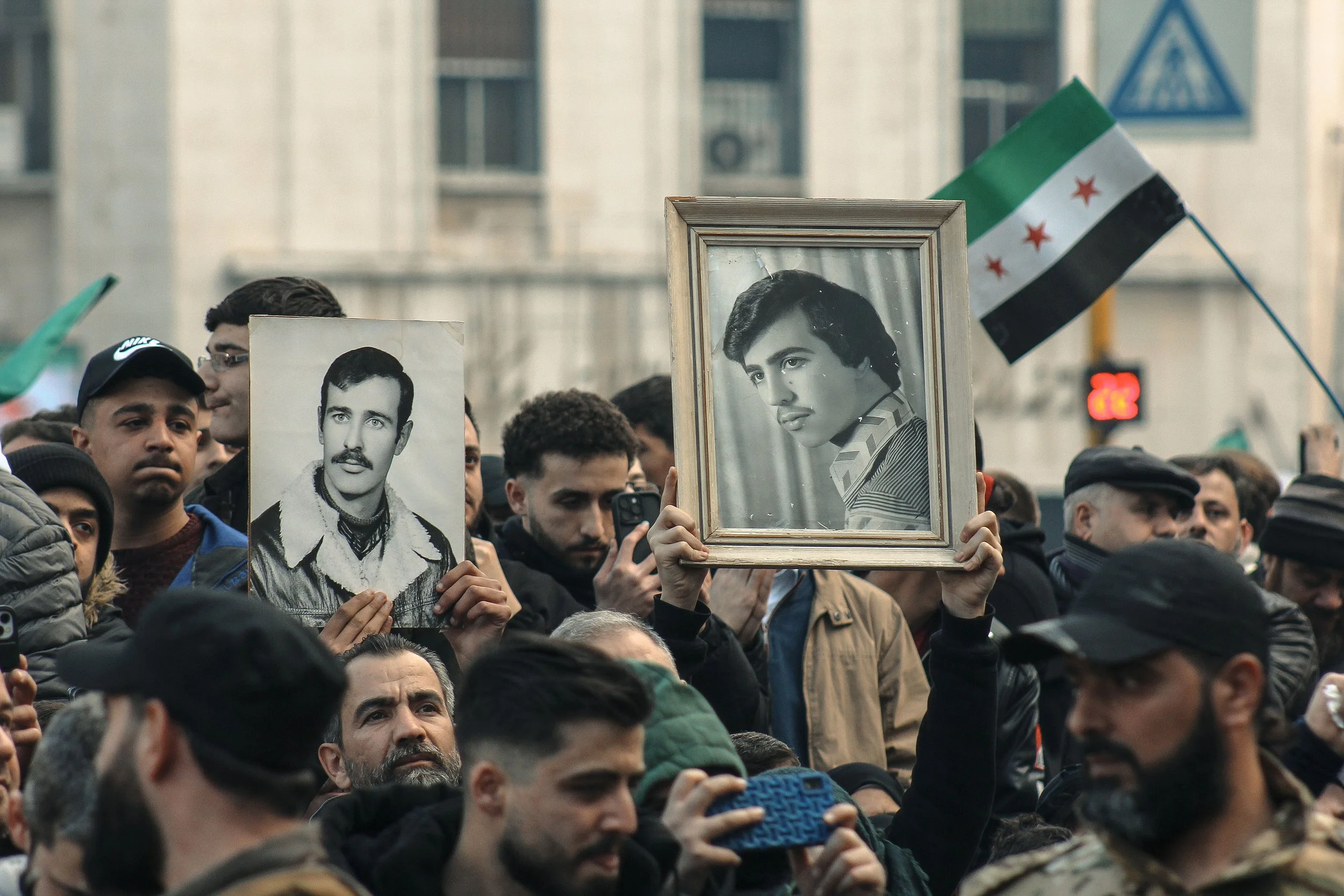Crowd of people holding black-and-white portraits of a man with dark hair, mustache, and a serious expression, along with a flag with green, white, black, and red stripes and three red stars, during a public gathering or demonstration.
