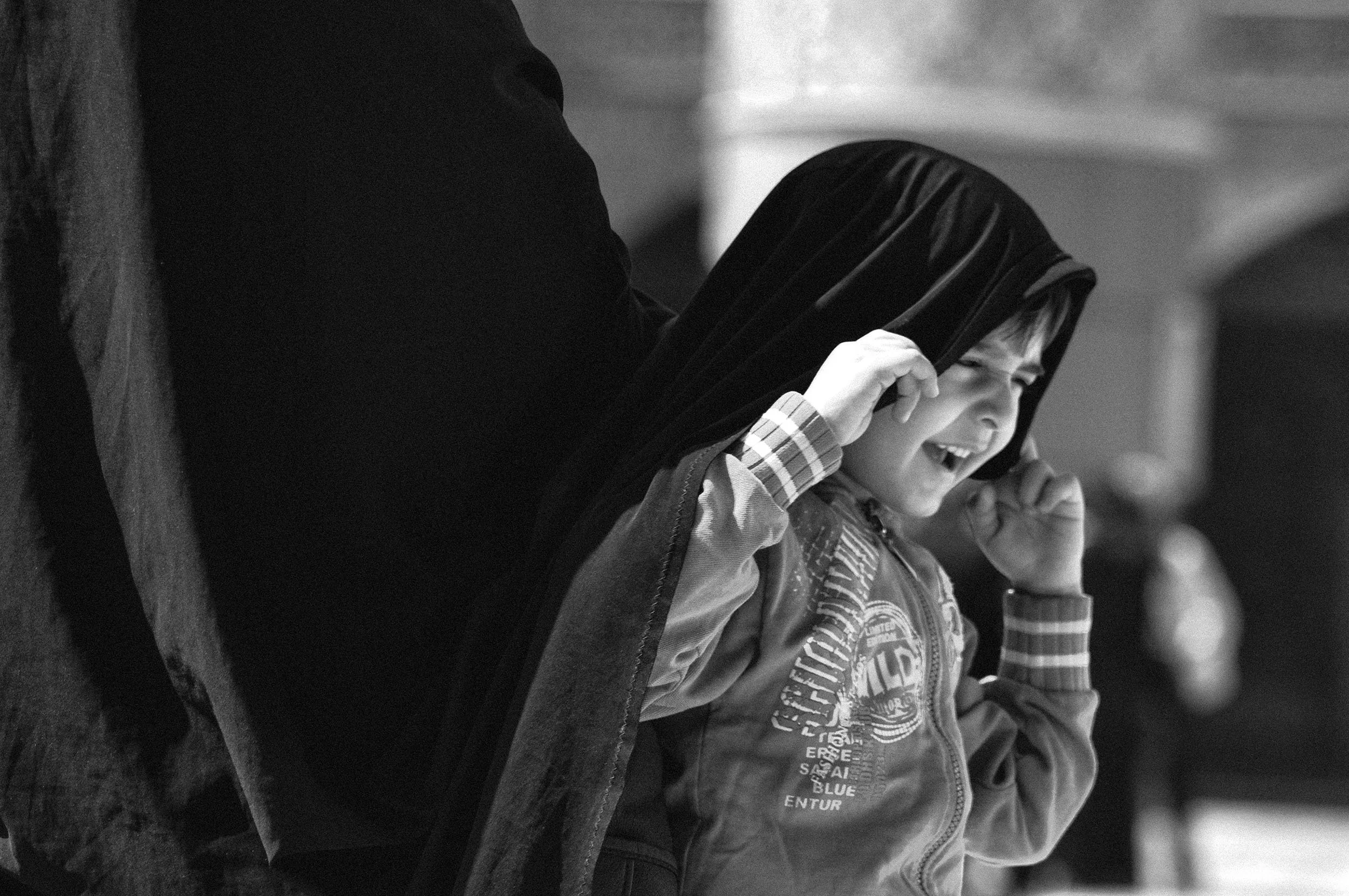 A young boy with a big smile, wearing a hoodie, is being hugged from behind by an adult, likely a woman, whose face is not visible. The boy appears happy and is holding the hood of his hoodie with both hands.