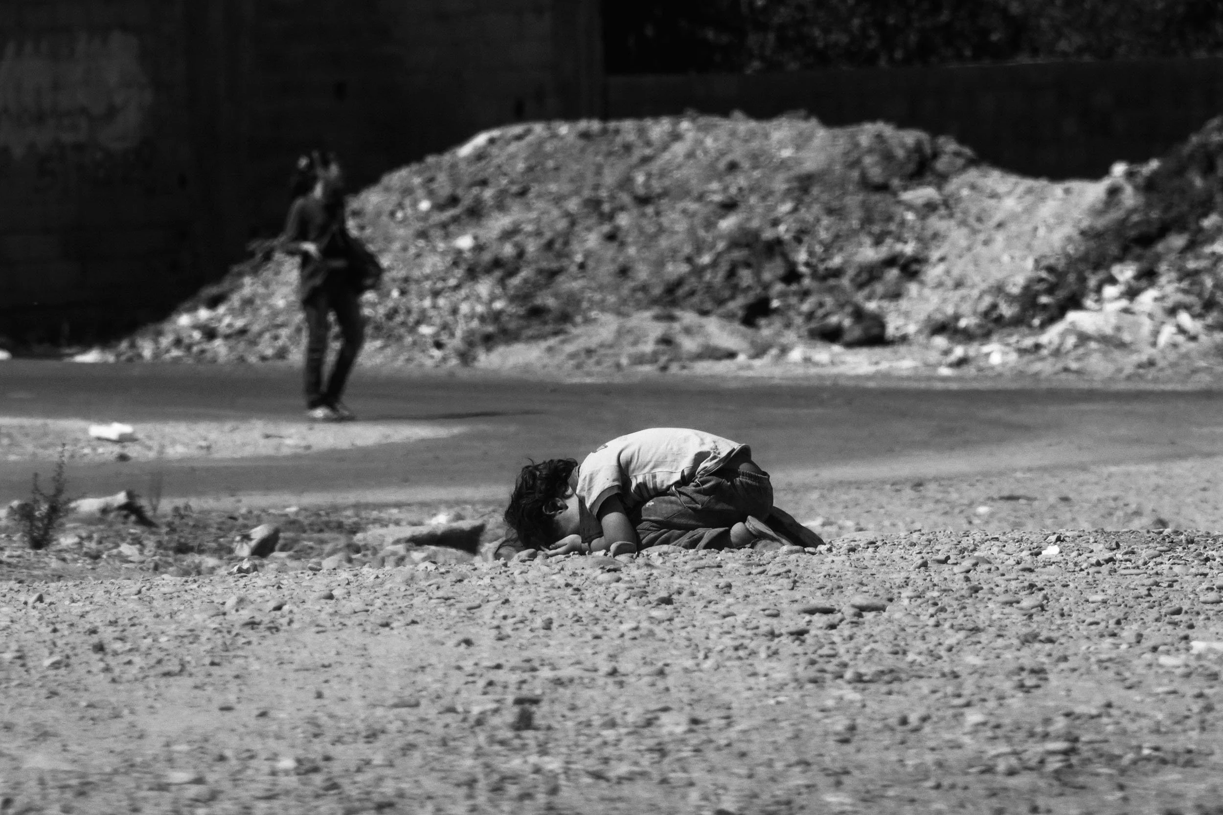 A young child kneels on the ground, bowing down with their head touching the dirt, in a desolate outdoor area with rocky terrain and another person walking in the background.