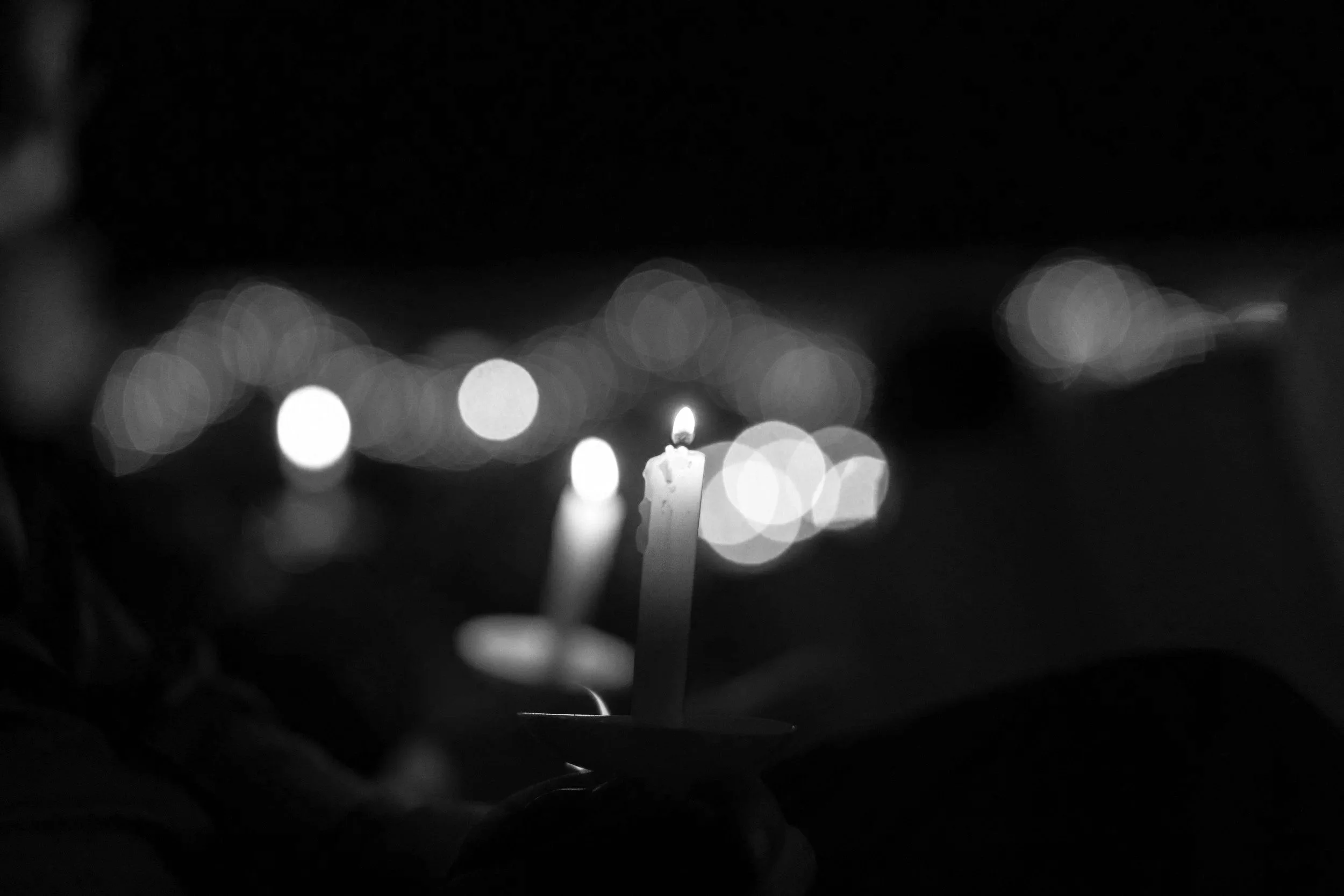 A hand holding a birthday candle with a lit flame, with blurred lights in the background.