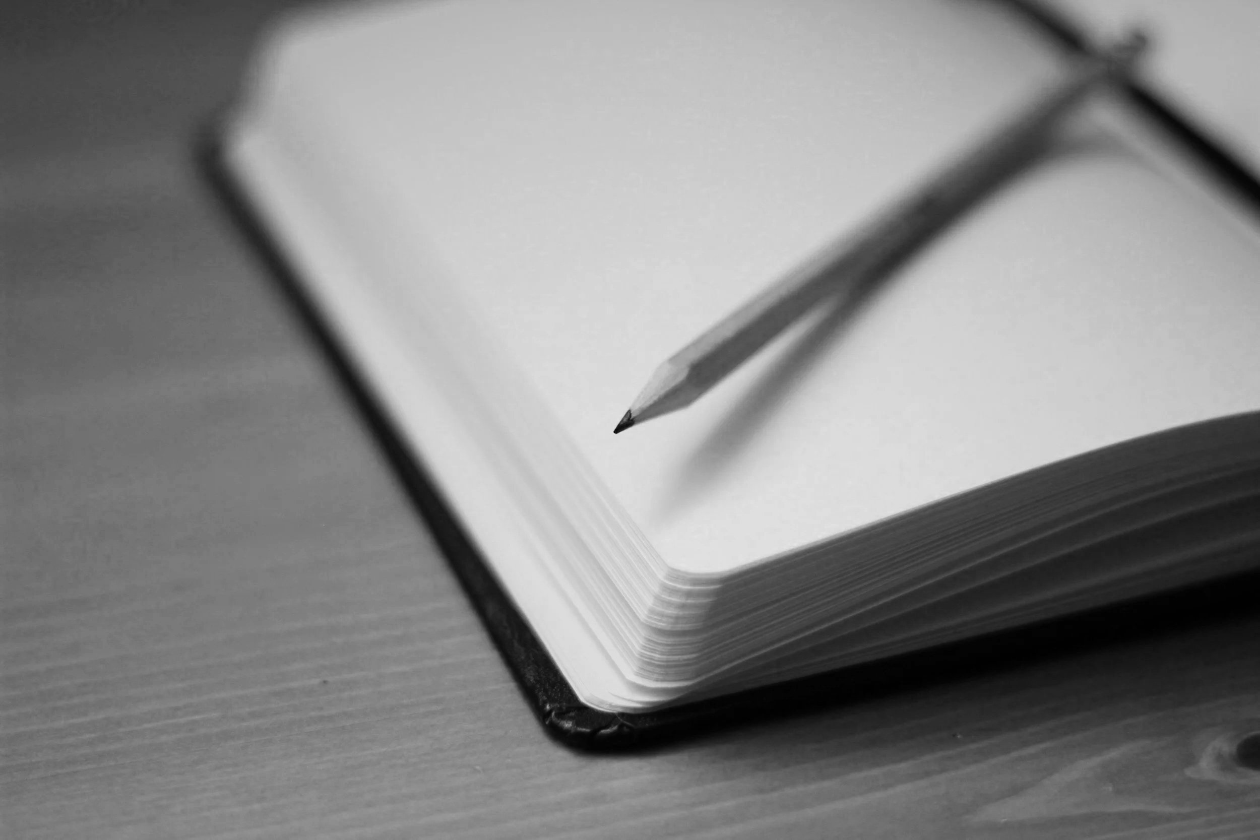 Black and white photo of an open notebook with a sharpened pencil resting on its blank page.