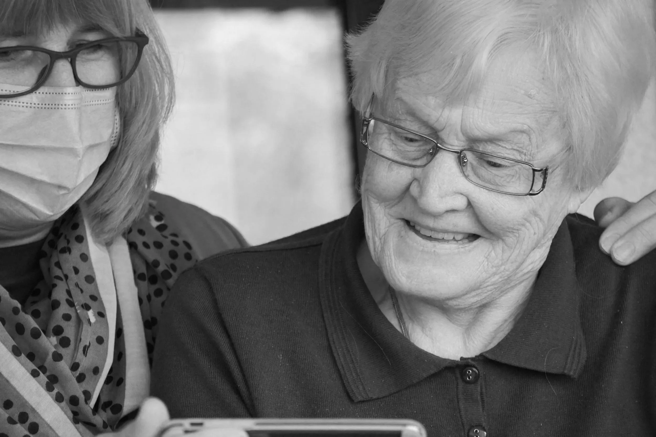 An elderly woman and a middle-aged woman, both wearing glasses, looking at a smartphone together. The elderly woman is smiling, and the middle-aged woman is wearing a face mask and a patterned scarf.