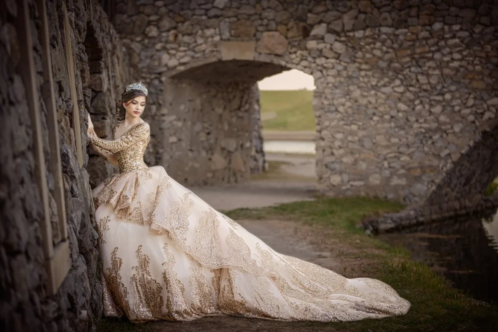 A Quinceañera in an elegant gold gown poses beside historic stone architecture, creating a timeless fairytale-inspired portrait. The dramatic dress and regal pose evoke the beauty and elegance of a modern princess.