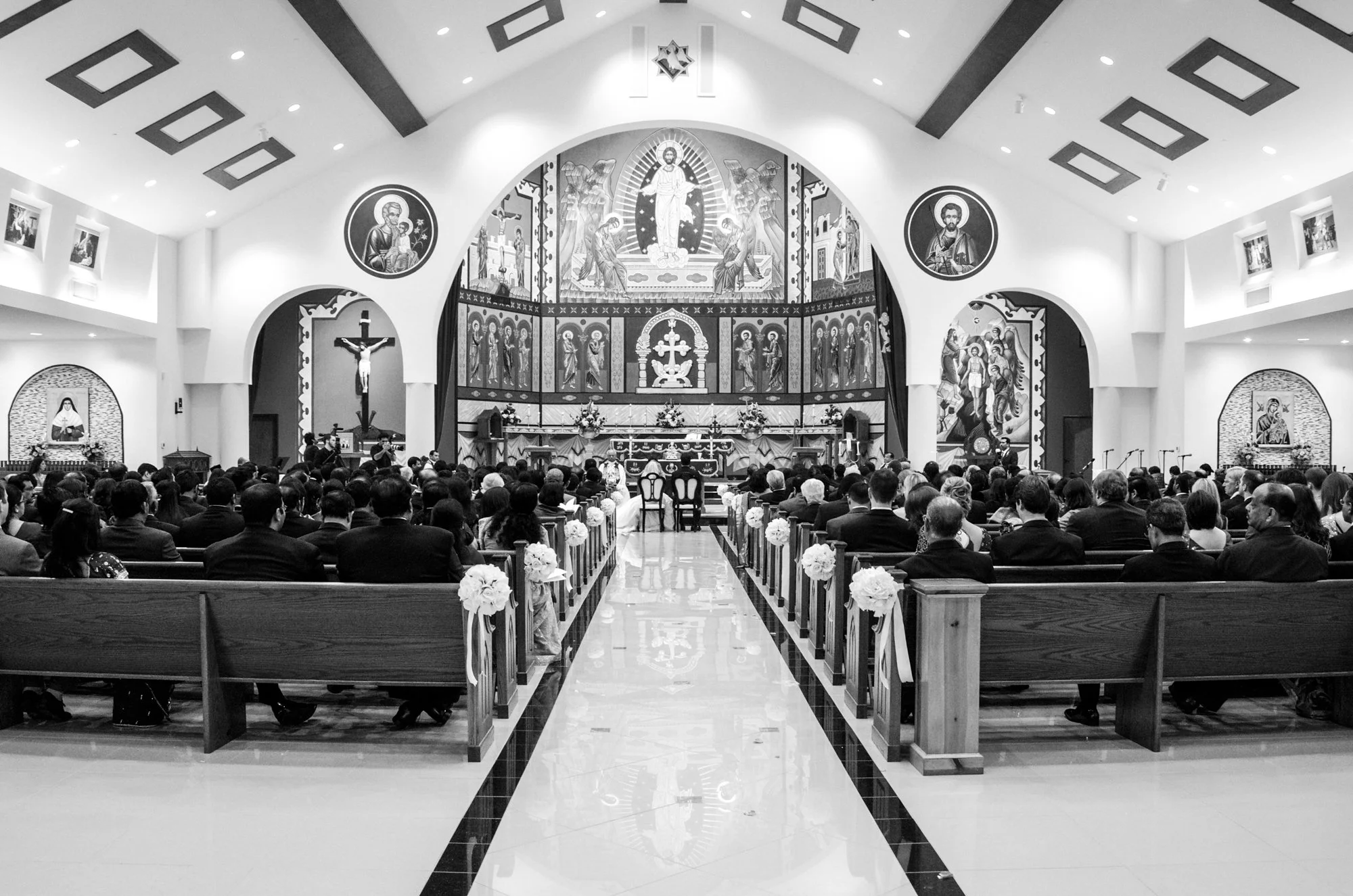A black-and-white wide-angle view of a church interior during a wedding ceremony, featuring rows of seated guests, ornate religious artwork, and a symmetrical aisle leading to the altar.