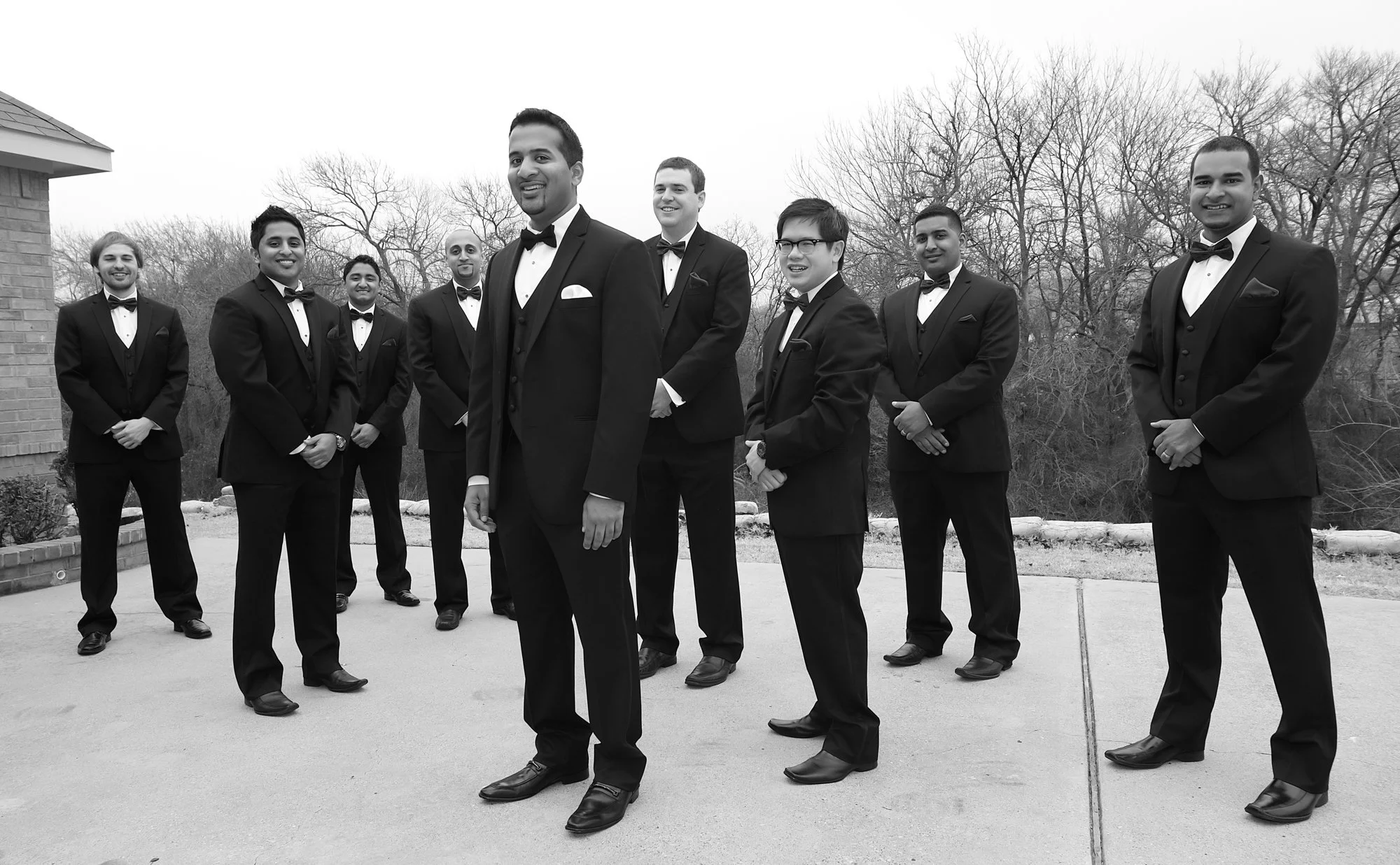 A black-and-white group portrait of the groom and his groomsmen standing outdoors in formal tuxedos, smiling confidently with bare winter trees in the background.