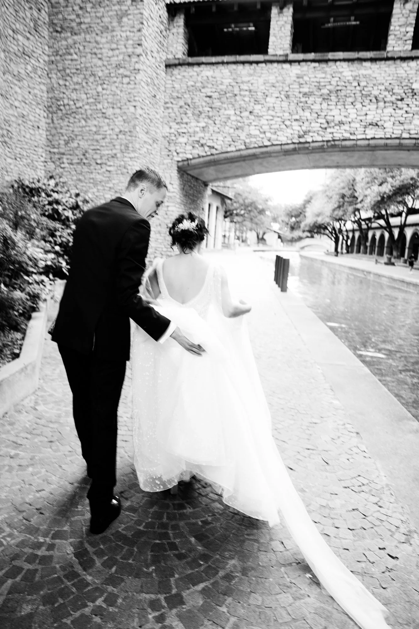 Romantic outdoor wedding portrait of the bride and groom walking hand in hand along a scenic stone pathway near water, captured from behind in black and white.
