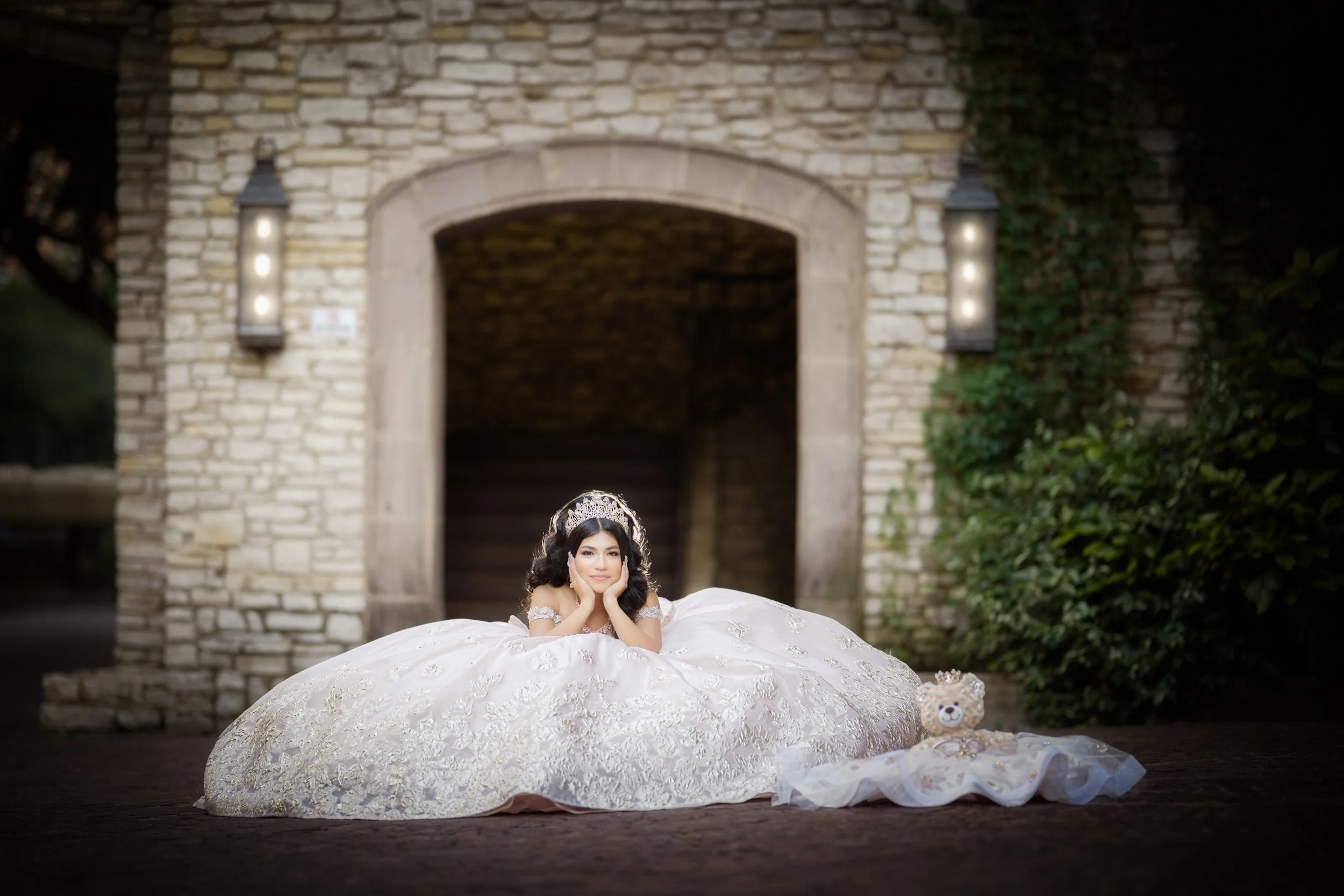 The quinceañera lies gracefully on the ground in front of a stone archway, resting her chin in her hands atop her gown. The expansive skirt creates a dramatic silhouette, while a small teddy bear accessory beside her adds a sentimental touch, blendin