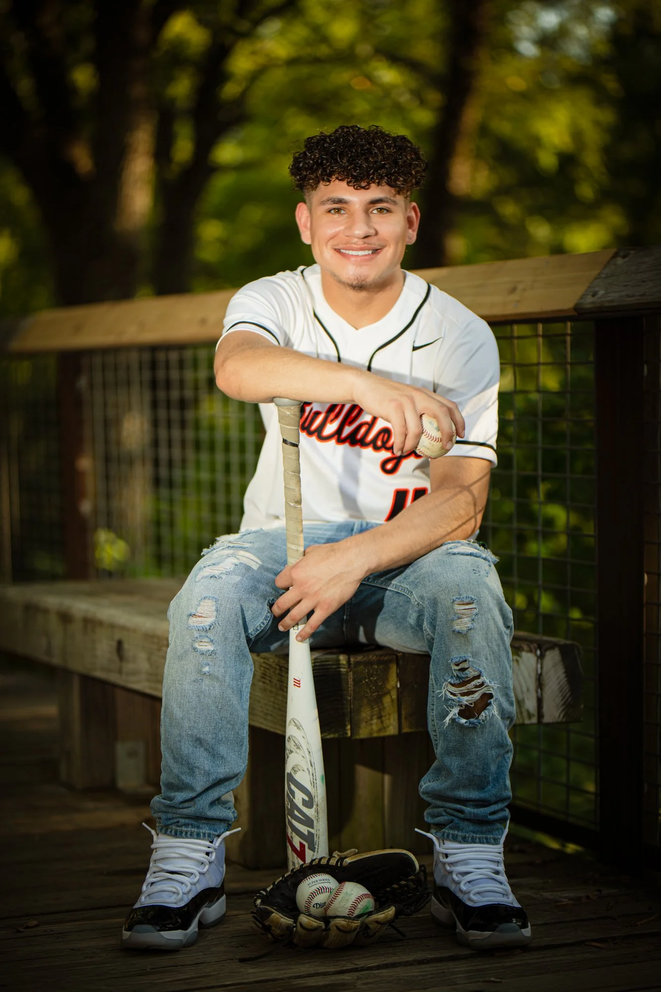 A baseball senior portrait of a young man smiling while holding a bat, seated outdoors on a wooden bench with trees softly blurred in the background.