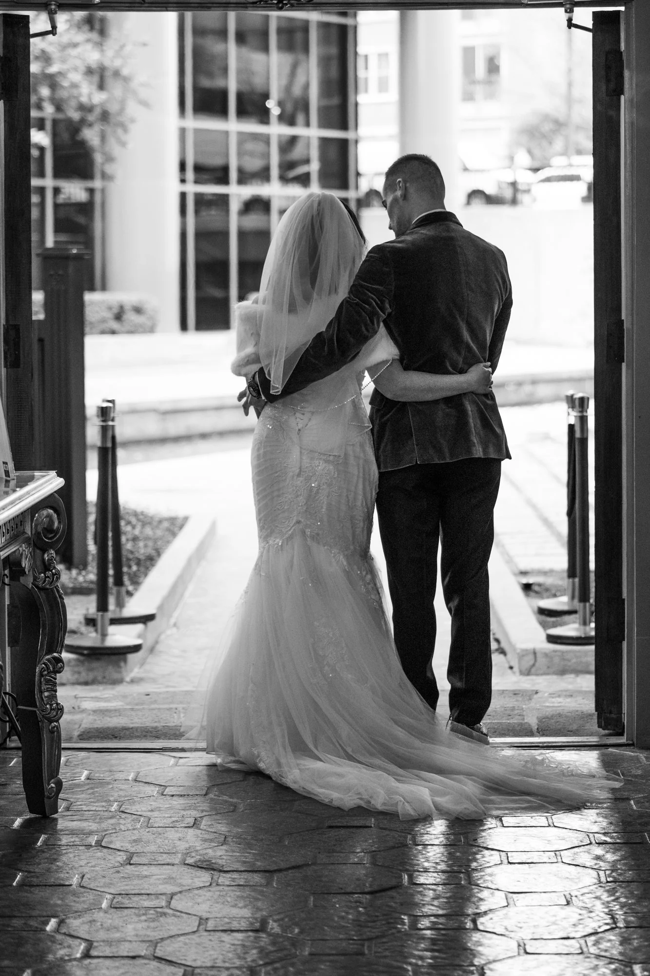 The bride and groom walk arm in arm toward the exit, seen from behind as her gown flows gracefully across the floor. This quiet, emotional scene symbolizes the beginning of their new journey together.