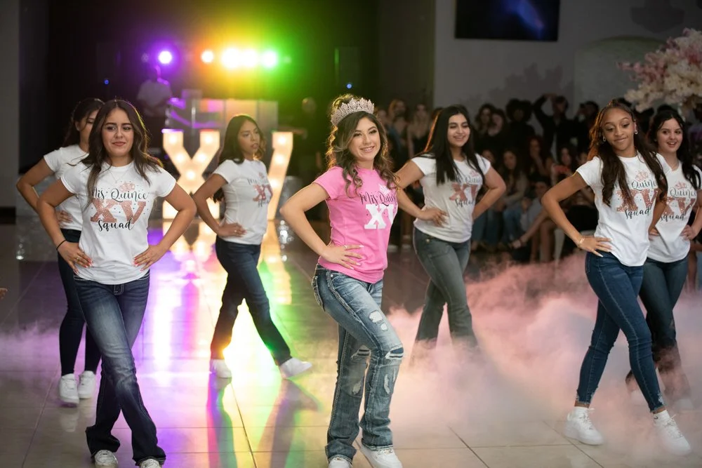 The Quinceañera leads her court during a choreographed surprise dance performance surrounded by colorful lighting and excited guests. This energetic image captures one of the most memorable moments of the celebration.