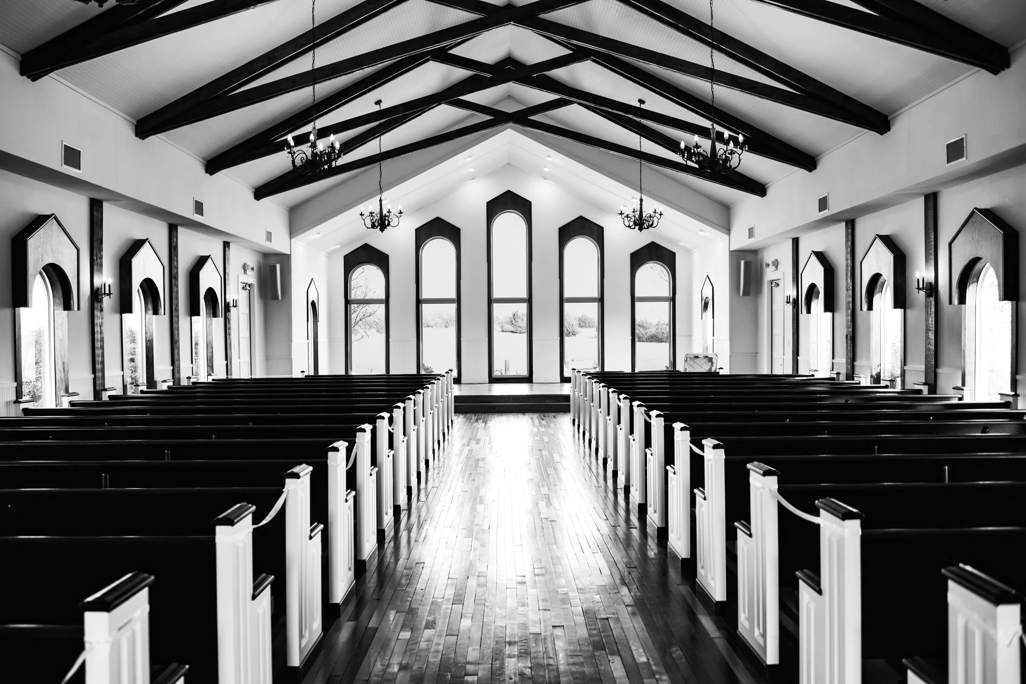 A black-and-white architectural interior shot of an empty chapel with wooden pews, vaulted ceilings, and tall arched windows, emphasizing symmetry, calm, and timeless beauty.