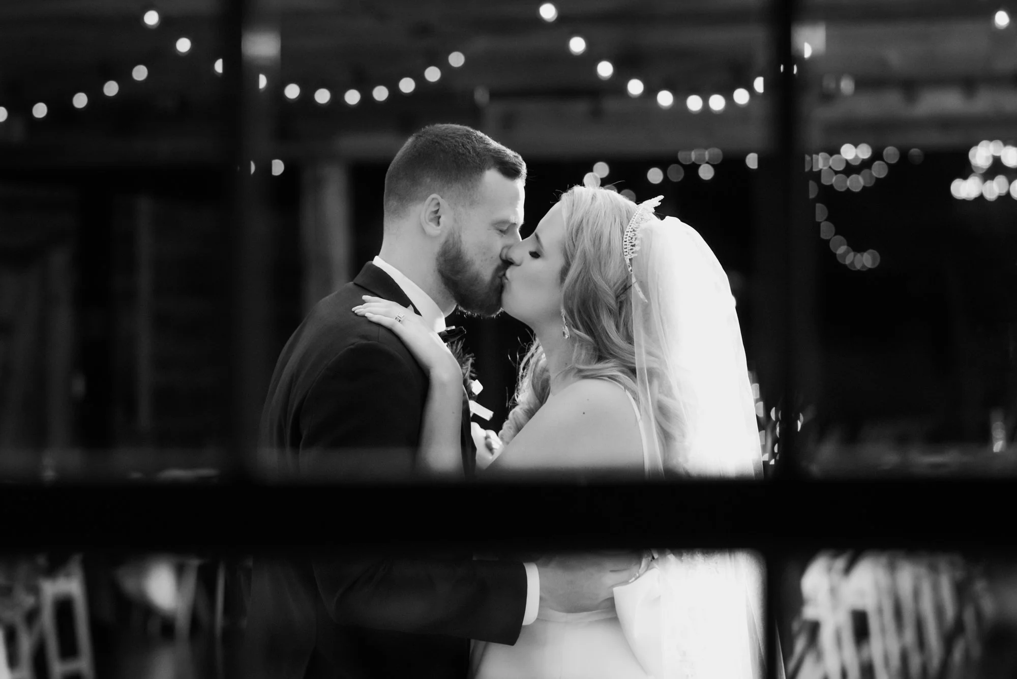 Romantic black and white close-up of a bride and groom sharing a kiss under glowing string lights during their reception, framed through architectural lines.