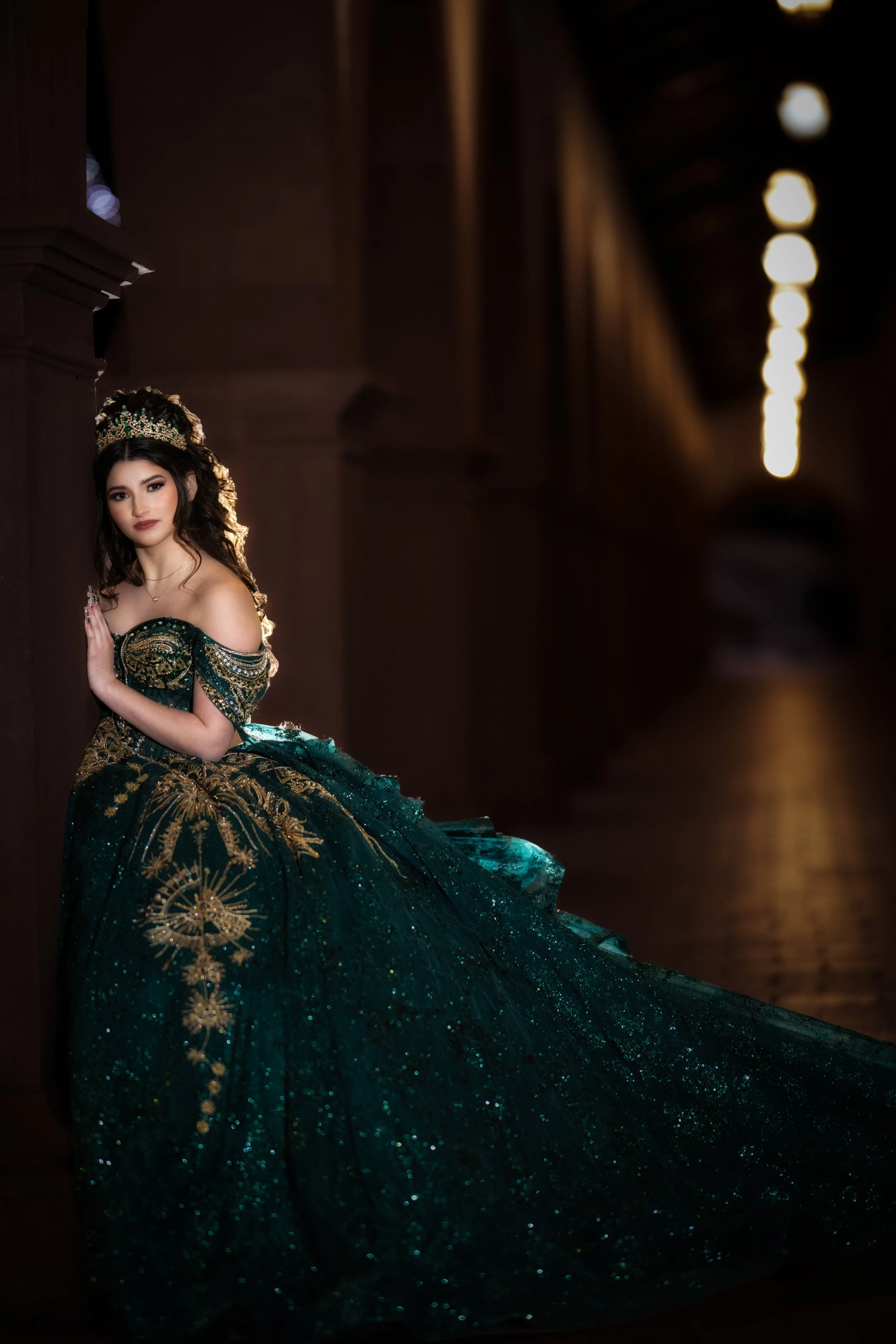 A dramatic Quinceañera portrait featuring a young woman in an emerald green ball gown with gold detailing, leaning against a stone column under warm architectural lighting that highlights elegance and confidence.